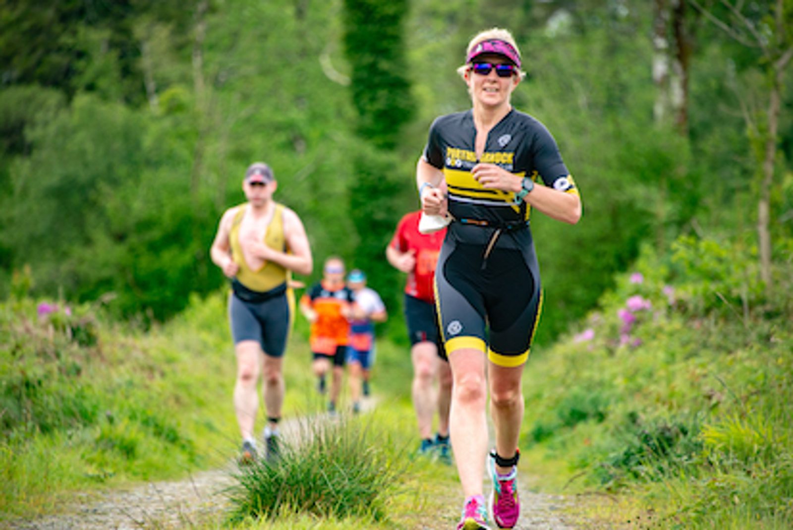 A group of people participating in a triathlon, running through a lush, green forest trail. The leading individual wears a black and yellow tri-suit, sunglasses, and a visor, smiling as they advance. Others follow behind, dressed in colorful athletic attire.