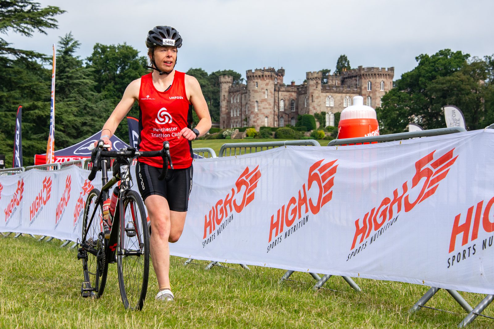A triathlete walks her bike along a grassy path at an outdoor event. She wears a red jersey with white text and a black helmet. A medieval-style castle and trees are in the background. The event is sponsored by High5, as indicated by the banners along the route.