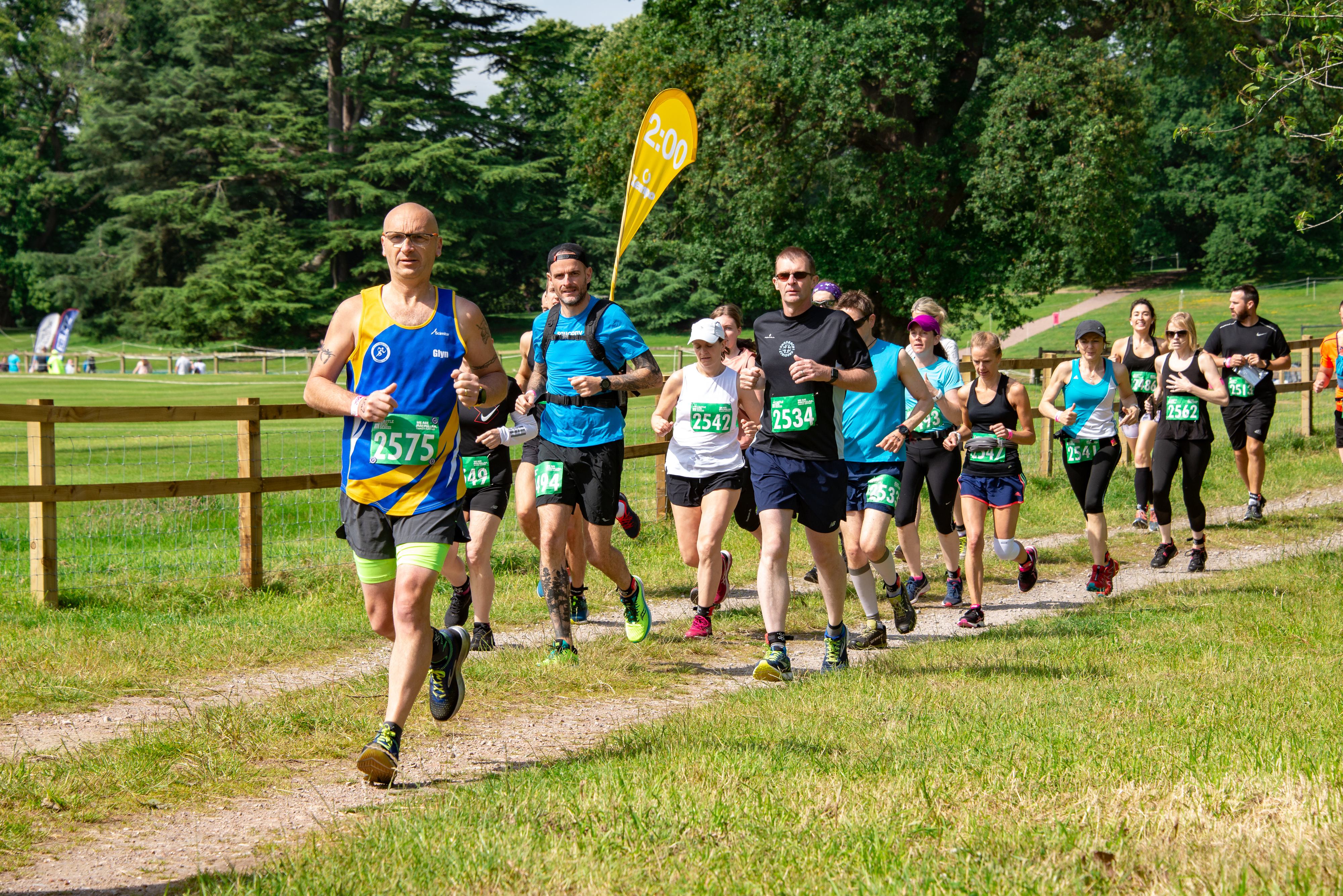 A group of runners, wearing numbered bibs, is participating in a race on a grassy trail through a park with trees in the background. The runners, in colorful athletic gear, are at various paces. Wooden fencing and a yellow marker flag can be seen alongside the trail.