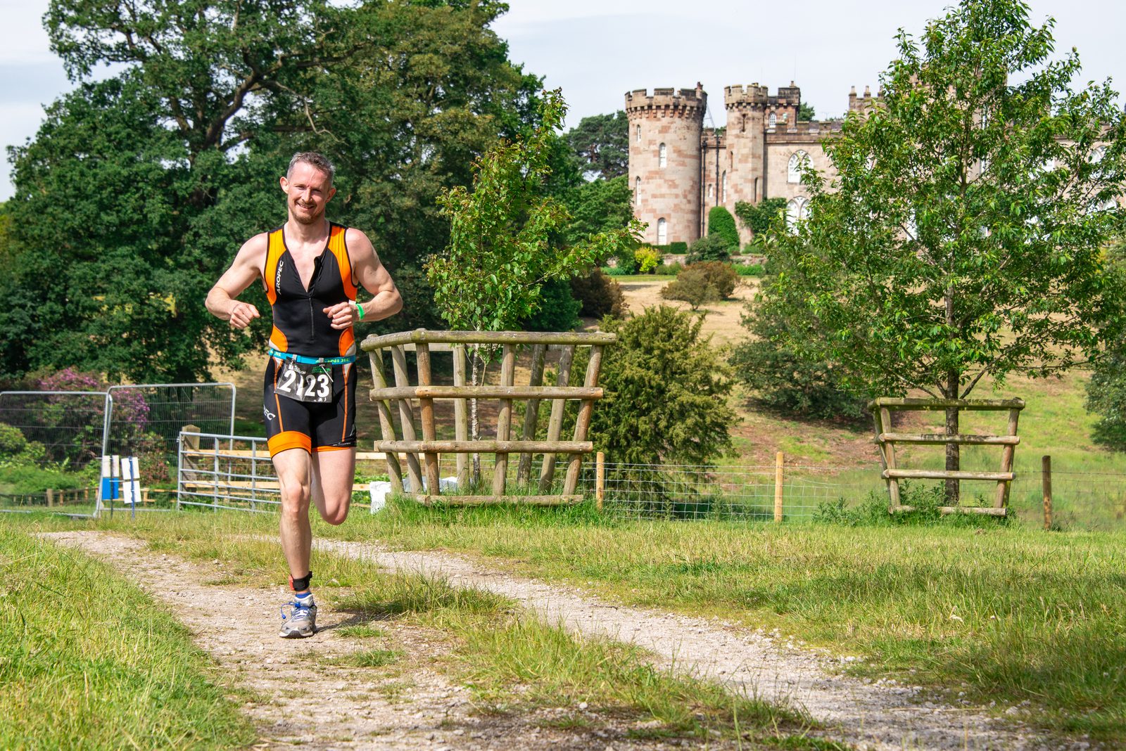 A man in athletic gear, including a bib with the number 123, runs on a dirt path through a grassy area with trees and wooden fences. A historic stone castle with turrets stands in the background under a partly cloudy sky.
