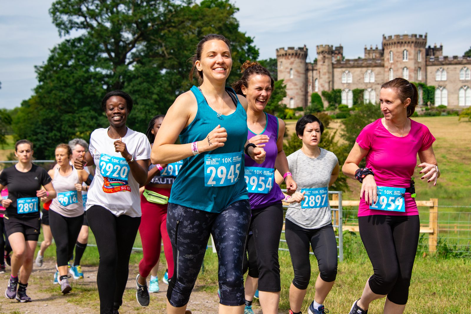 A group of runners, mostly women, participate in a race with numbered bibs on their shirts. They are jogging on a grassy path with a castle-like building in the background. The runners appear cheerful and energetic under a sunny sky.