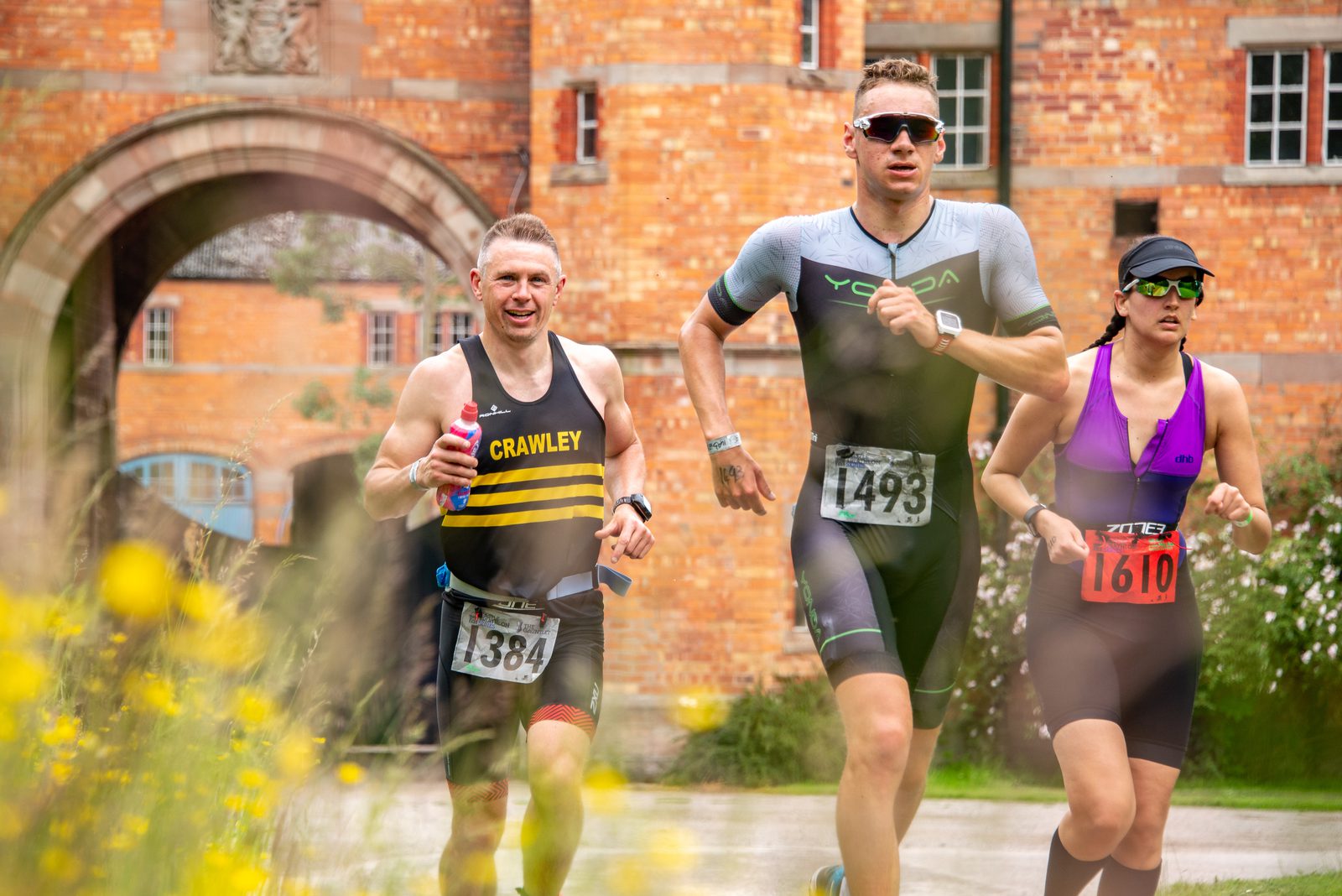 Three athletes participate in an outdoor race, with bib numbers 1384, 1493, and 1610. They run past a historic brick building with an archway. The foreground features yellow wildflowers, adding color to the scene. All athletes are wearing athletic gear and sunglasses.
