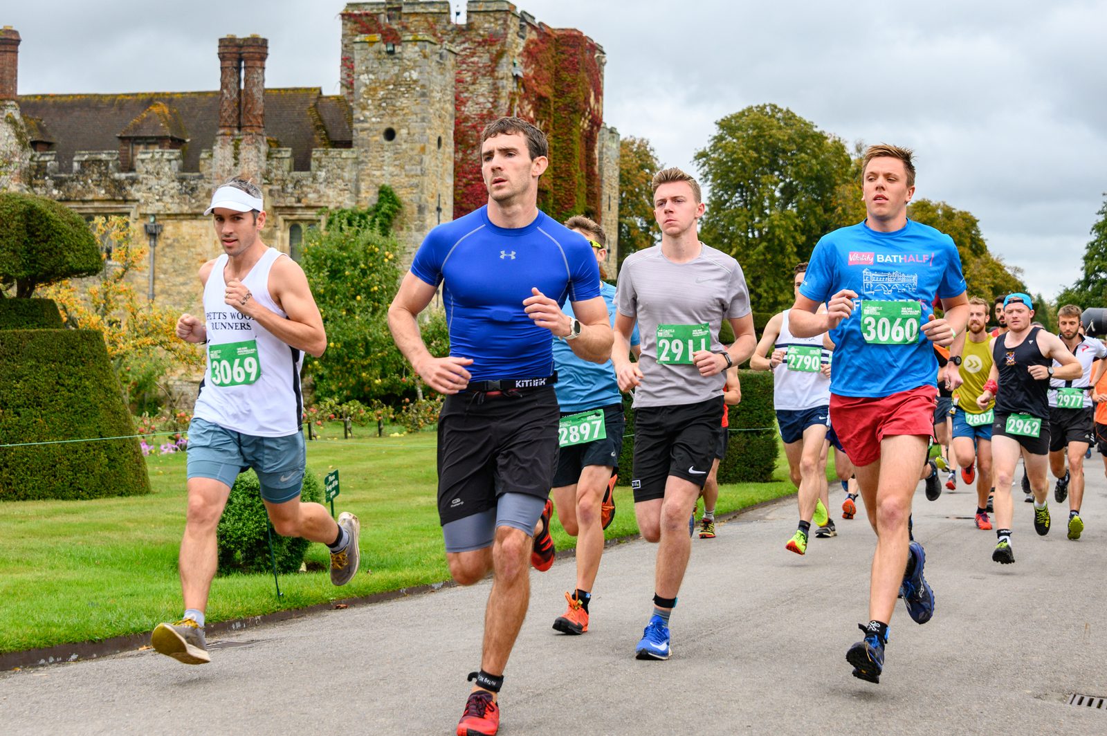 A group of runners participating in a marathon race on a paved path, with an ancient stone building and well-maintained gardens in the background. The runners wear various athletic outfits and numbered bibs, indicating their participation in the race.