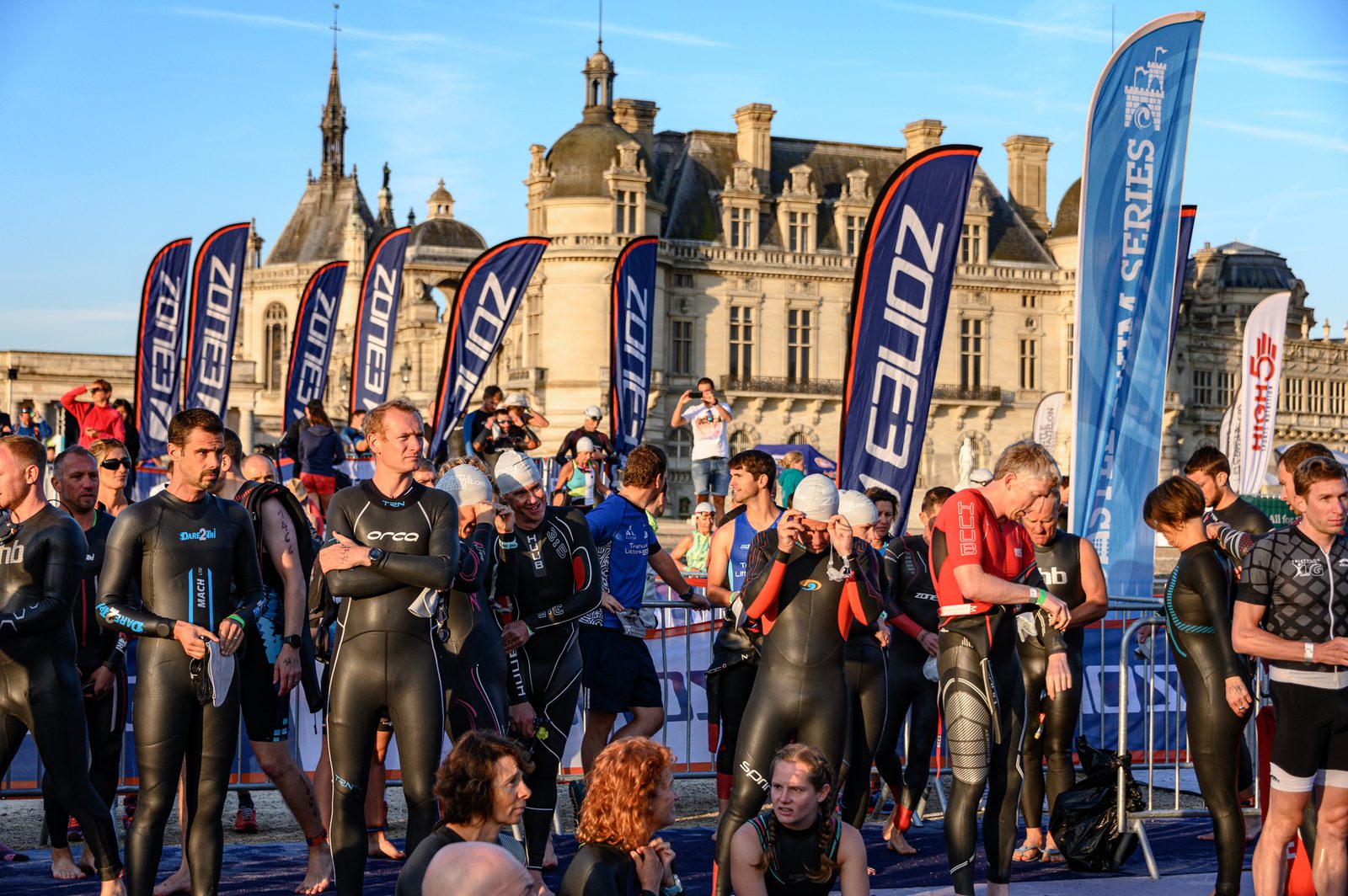 A crowd of athletes in wetsuits gathers at the starting line of a triathlon event. They are preparing for the swim leg of the race, with a historic building and several race banners in the background. Some athletes are adjusting their gear and chatting among themselves.