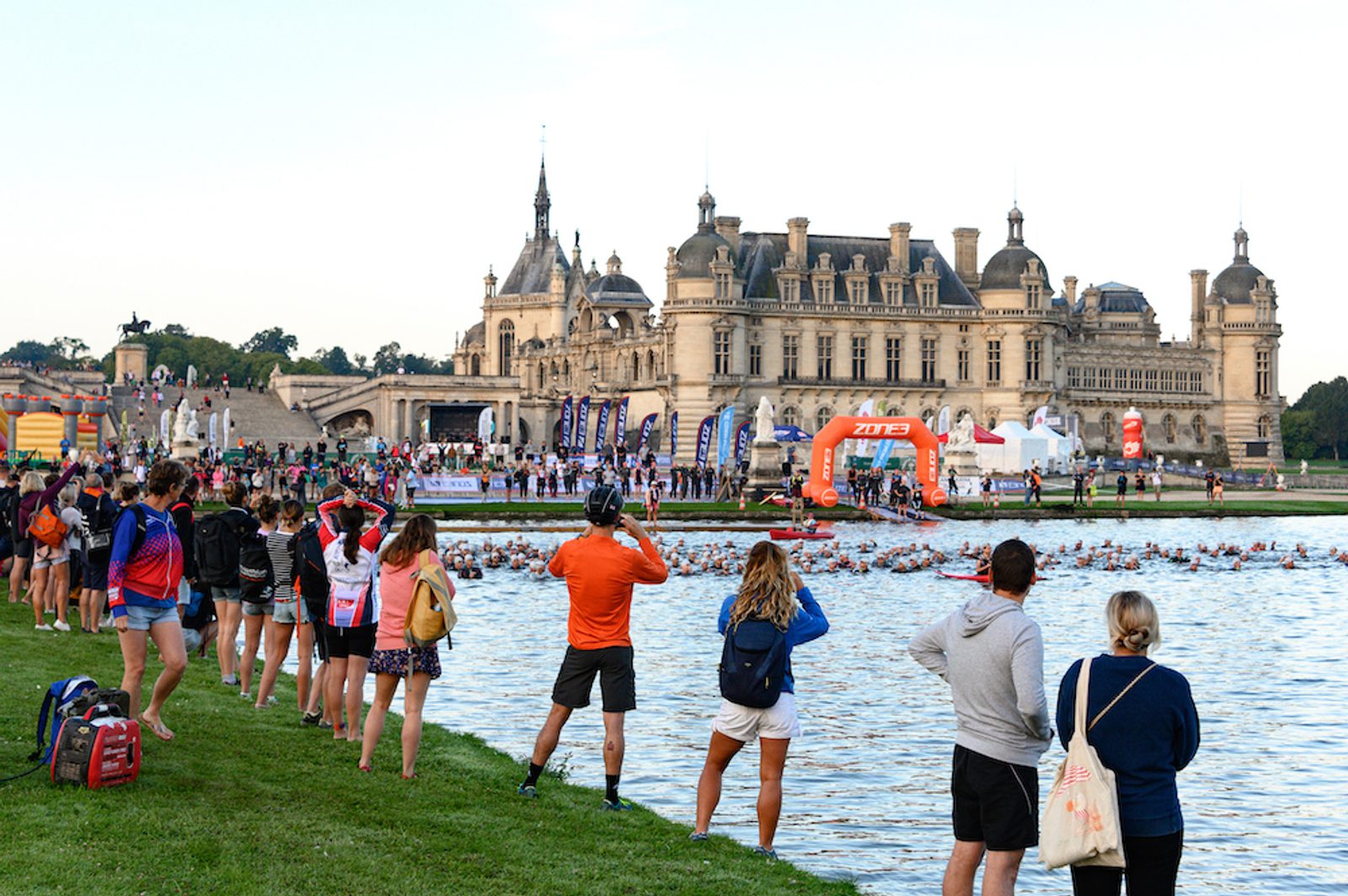 Spectators line the bank of a waterway, watching participants in a race swim past. A grand chateau is visible in the background, with flags and tents set up near the water's edge, creating a festive atmosphere.