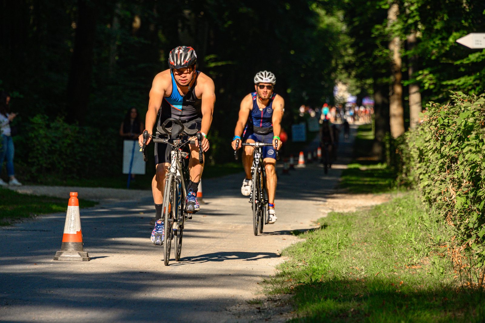 Two cyclists in full gear are racing on a shaded forest road during a sunny day. An orange and white traffic cone marks the path. Spectators and more cyclists are visible in the background. The road is flanked by green foliage and trees.