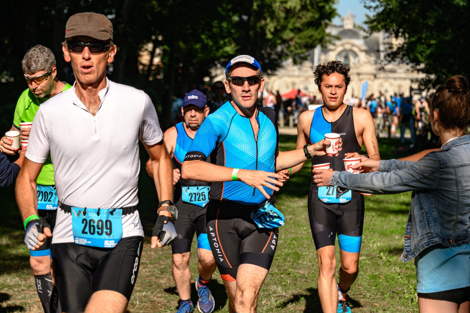 A group of triathletes in blue and black gear run on a grassy path during a race. One racer in the foreground, wearing sunglasses and a white shirt, reaches for a cup from a volunteer. Other athletes and trees are visible in the background.