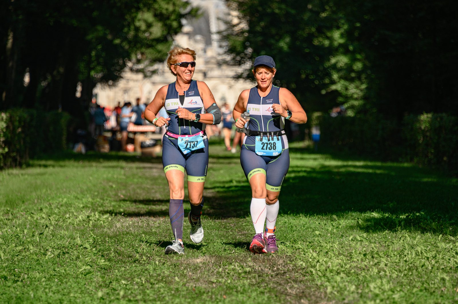 Two women wearing athletic gear (matching navy blue tops and shorts) participate in a race, running side by side on a grassy path. Both are smiling and wearing sunglasses. Other runners and a wooded area with sunlight filtering through the trees are seen in the background.