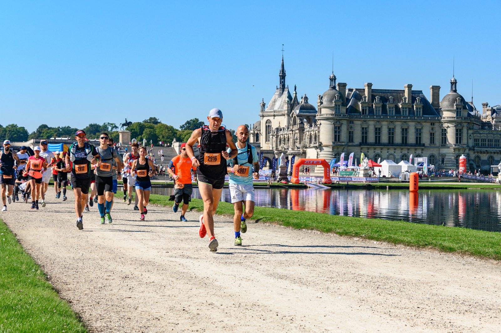 A group of runners participates in a race on a sunny day, with a historic chateau in the background. The course runs alongside a body of water, and the runners wear various athletic gear and race bibs. The scenery includes green grass and a blue sky.