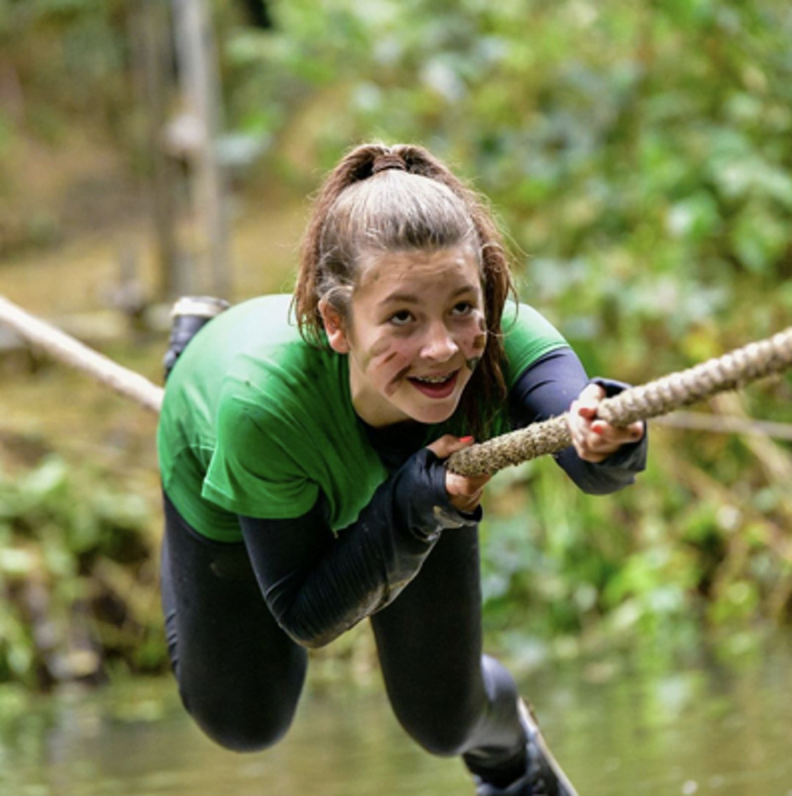A young person with long hair in a ponytail, wearing a green shirt and black pants, crawls on a rope bridge over water. They have a determined look on their face and camouflage paint on their cheeks. The background is blurred foliage.