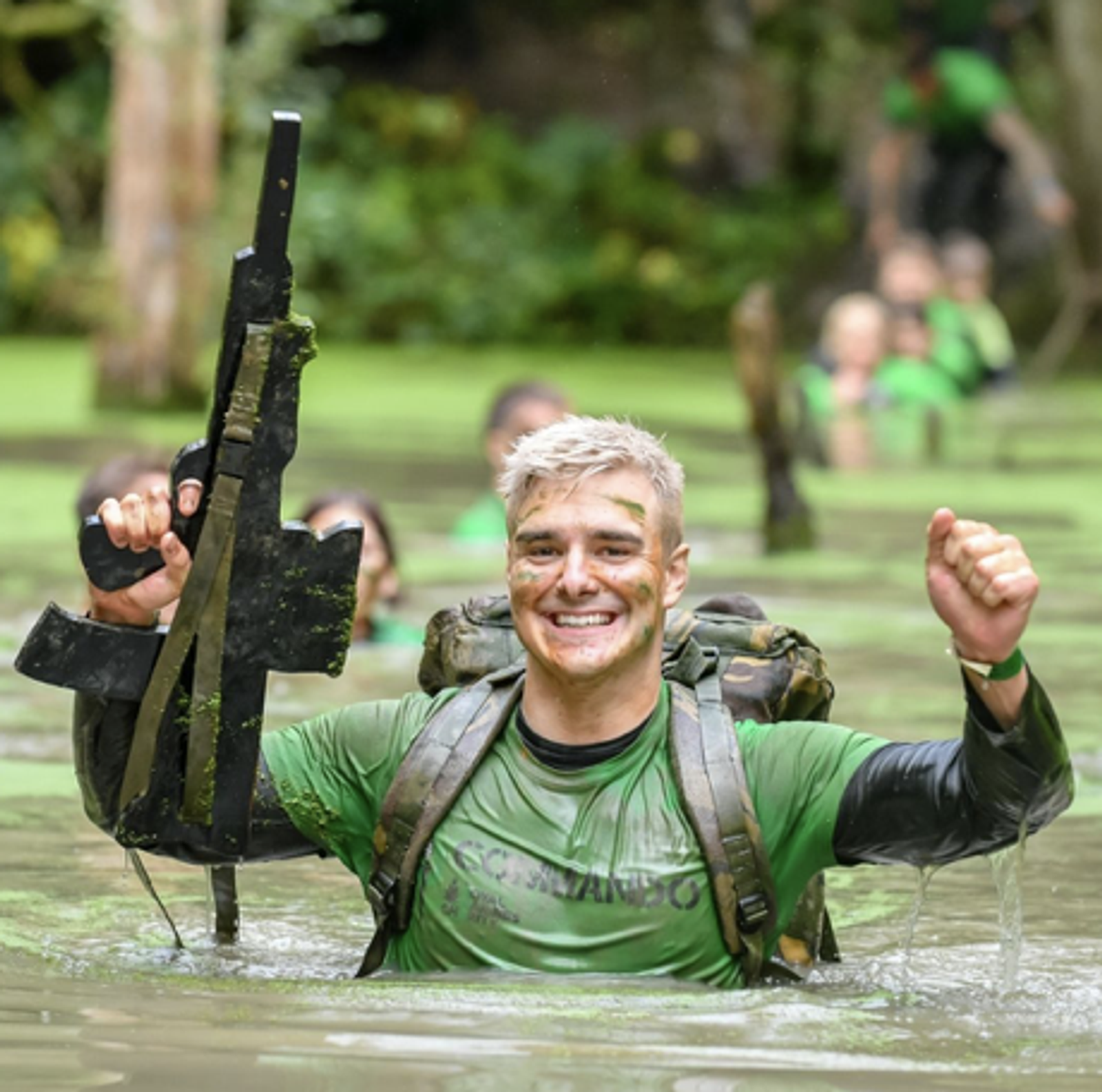 A man in military attire, with a green shirt and face paint, jubilantly raises a weapon with one hand while wading through a swamp with water up to his chest. He has a big smile on his face and other individuals in similar gear can be seen in the background.