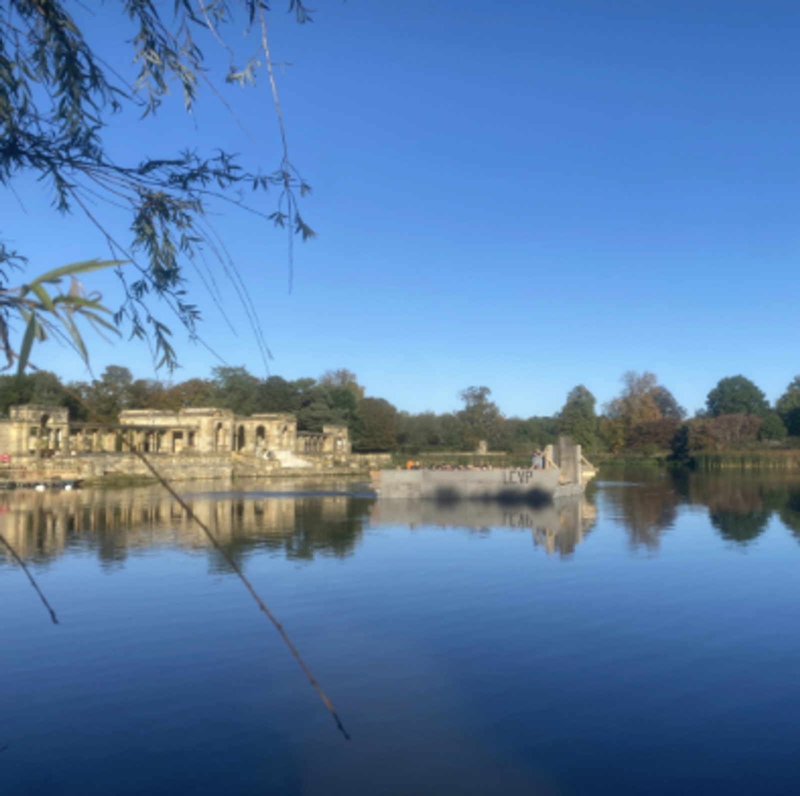 A serene body of water reflects the blue sky above and the surrounding landscape. Ancient stone ruins and lush green trees are visible on the opposite shore. Drooping branches frame the scene, adding to the tranquil atmosphere.