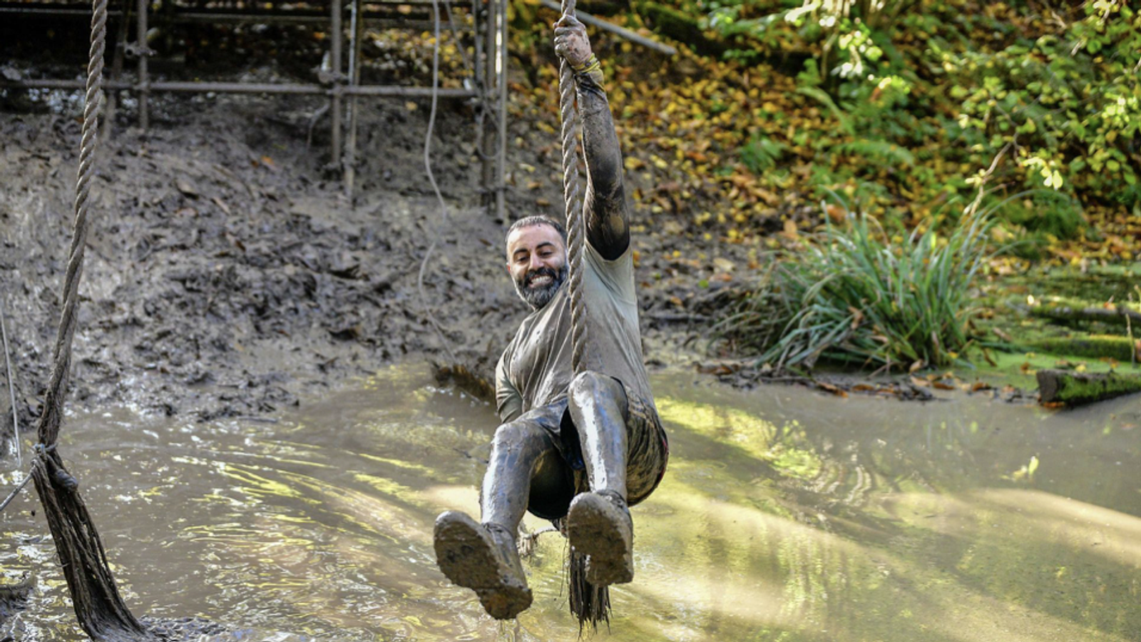 A man swings from a muddy rope over a swampy area with a big smile on his face. The surroundings are filled with greenery and fallen leaves. He is covered in mud from head to toe, indicating he is participating in an outdoor adventure or obstacle course.