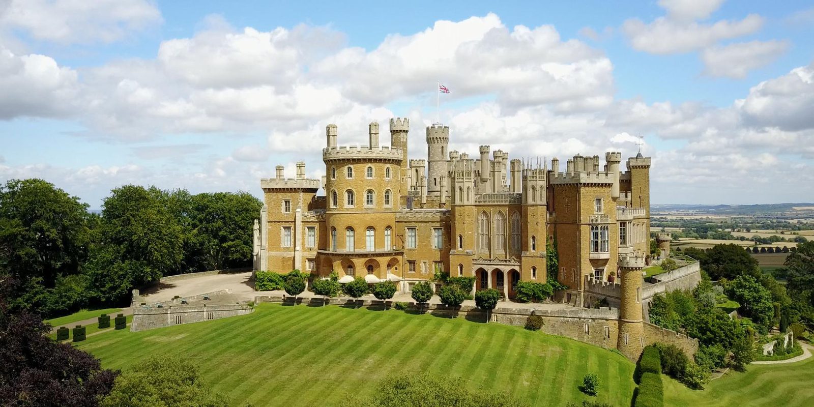A scenic image of a large, historic castle set amidst well-manicured lawns and lush greenery. The castle has multiple towers and turrets with an English flag waving on top. The sky is partly cloudy, and the surrounding landscape is expansive and picturesque.