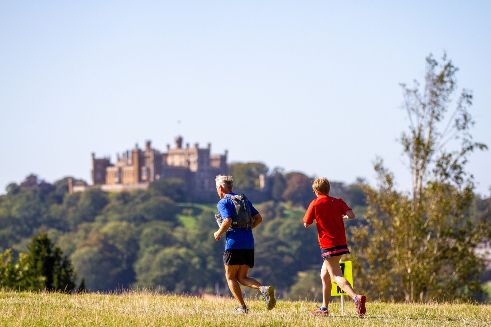 Two people jog on a grassy field with a historical castle set on a distant hill in the background. The sky is clear and blue, and a tree stands to the right side of the image. The jogger on the left is wearing blue, and the one on the right is in red.