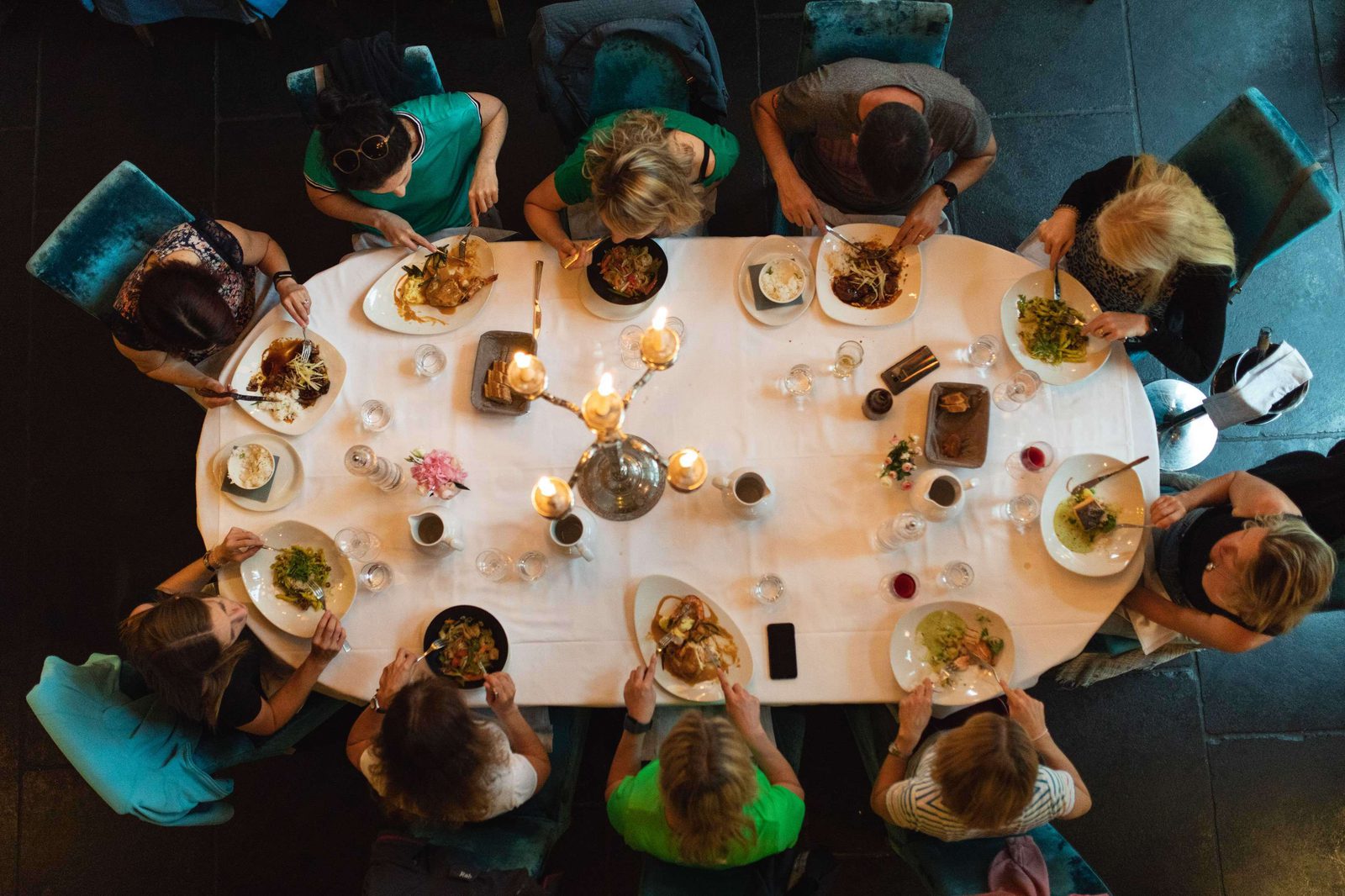 A group of people are seated around a large rectangular dining table, viewed from above. The table is set with various dishes and lit with decorative candles. Plates full of food are in front of each person, and they appear to be enjoying a meal together.