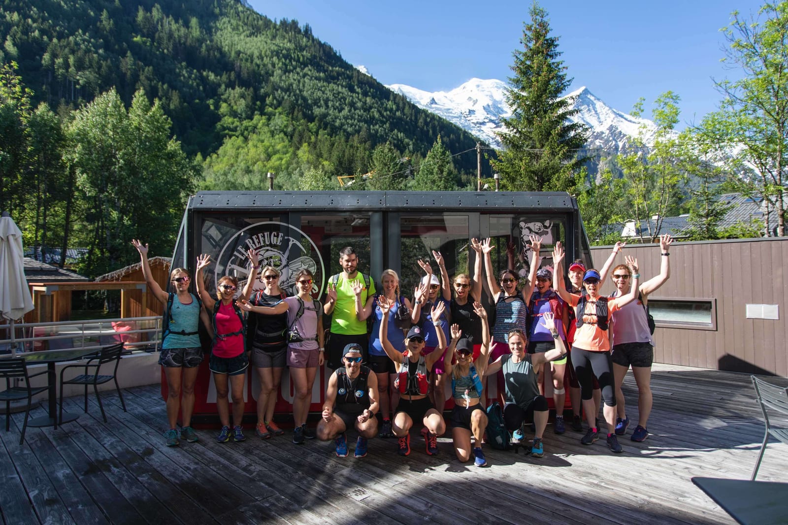 A group of people stands on a wooden deck with their hands raised, smiling for the camera. The group is dressed in athletic clothing, with a mountainous, forested landscape and a cable car in the background. The sky is clear and blue.