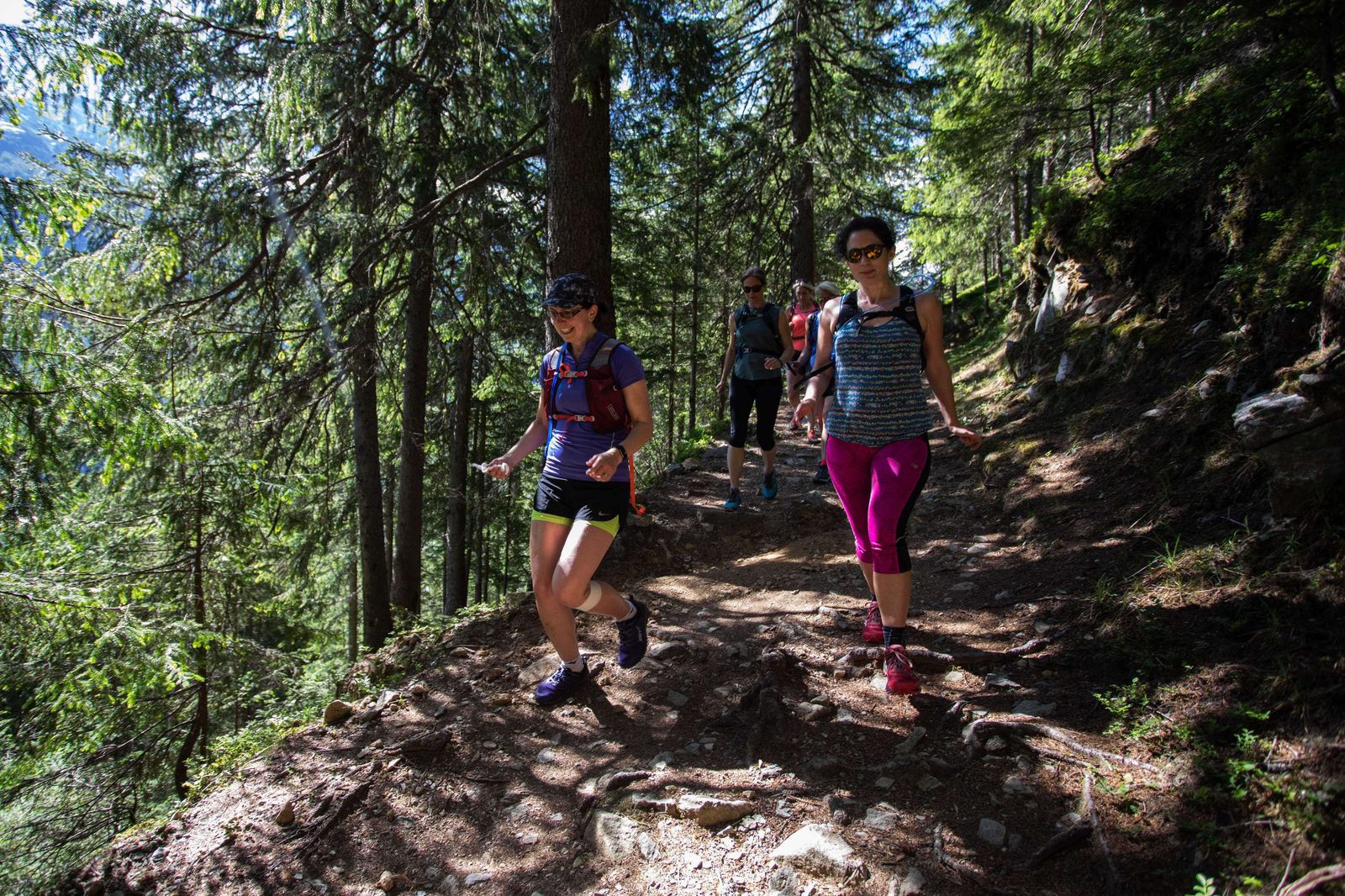A group of hikers walk along a forest trail, surrounded by tall, green trees. Two women in the foreground lead the way, wearing athletic clothing, backpacks, and sunglasses. Dappled sunlight filters through the canopy, casting shadows on the path.
