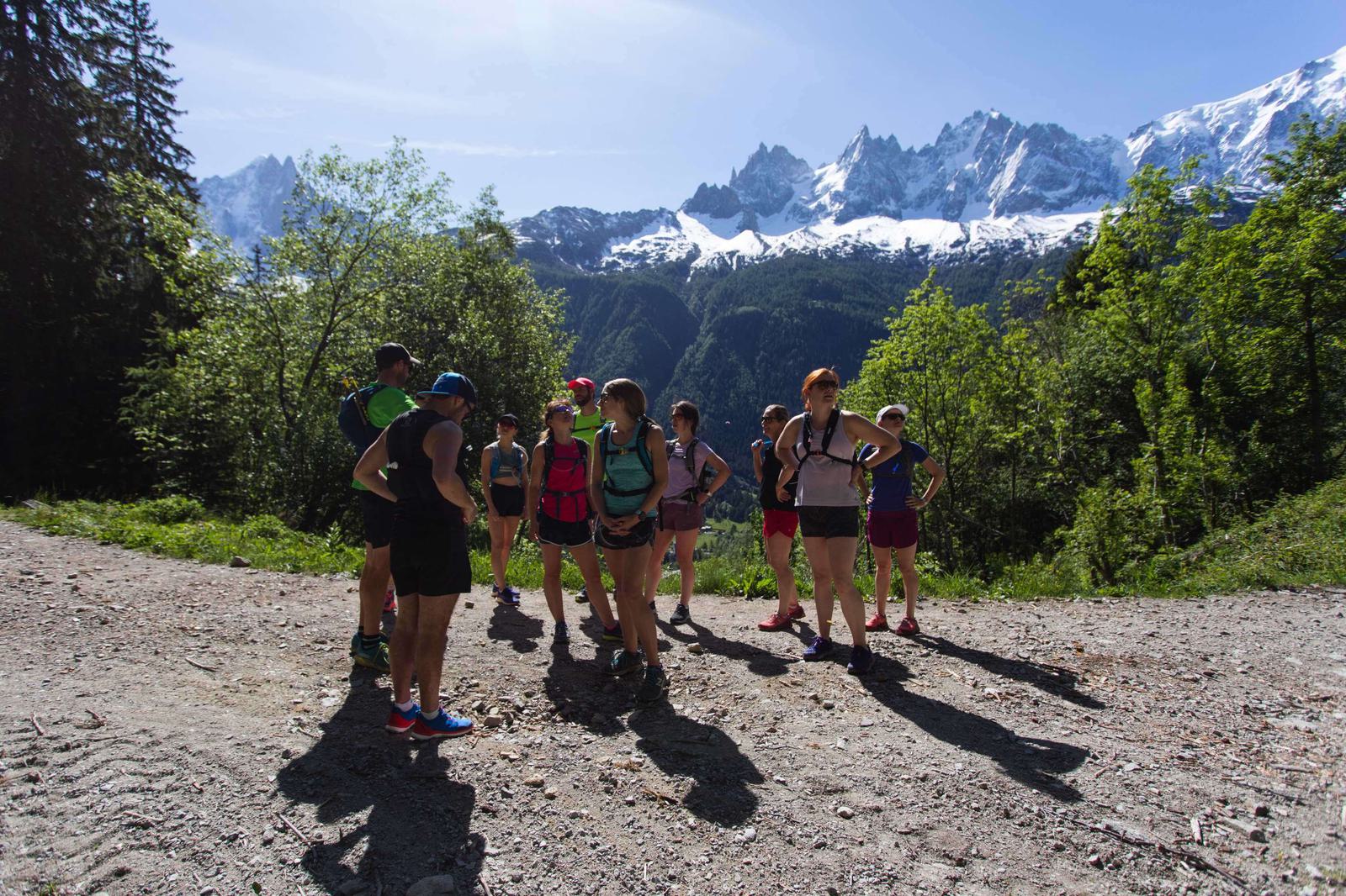 A group of hikers stands on a dirt path in a mountainous area. The sun casts long shadows, and everyone wears outdoor athletic gear and backpacks. Snow-capped mountains and lush green trees fill the background under a clear blue sky.