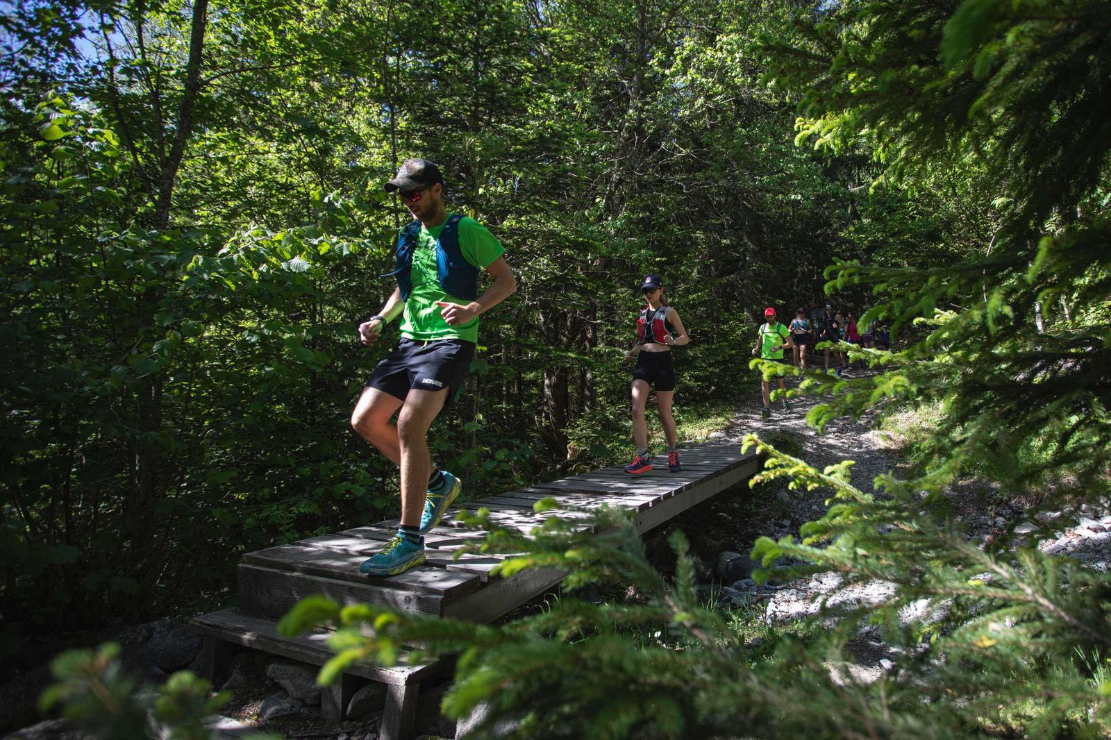 A group of runners cross a wooden bridge on a forest trail. The leading runner wears a green shirt and cap, while others follow behind on the path through the dense greenery. Sunlight filters through the trees, casting dappled light on the scene.