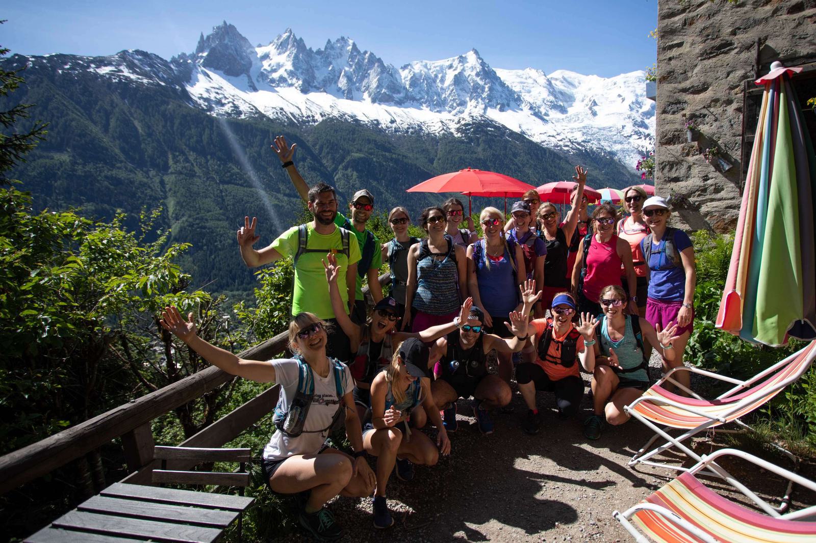A group of hikers, dressed in bright outdoor attire, pose cheerfully with raised hands at a scenic mountain viewpoint. Snow-capped peaks and lush green hills form the breathtaking background. Some lounge chairs and an umbrella are visible to the side.
