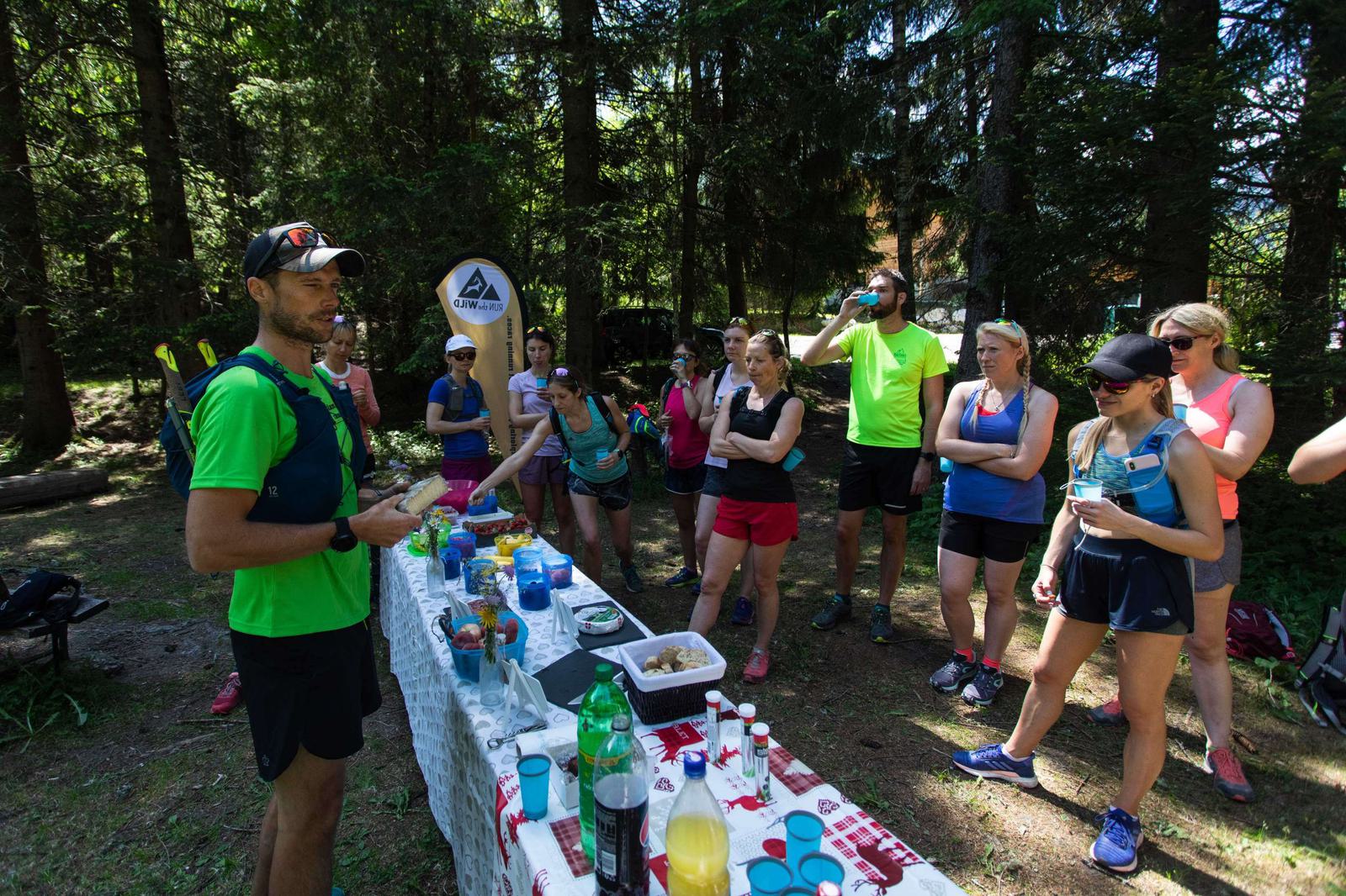 A group of runners in athletic gear stand around a long table set up outdoors in a forest. The table is covered with various drinks and snacks. One man, seemingly the instructor, is speaking to the group while they listen attentively.