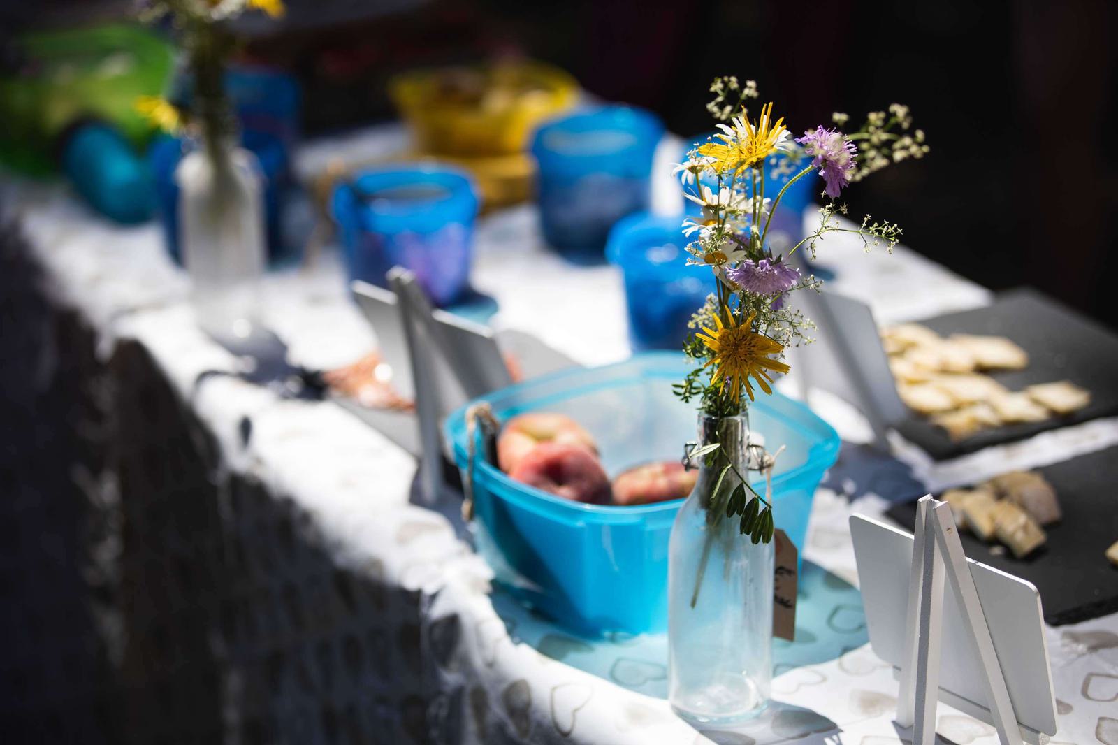 A sunny outdoor table with floral arrangements in small glass bottles, a light blue bowl containing apples, and blue cups. Plates of sliced food are on the table, and white signboards are displayed. The tablecloth has a subtle heart pattern.