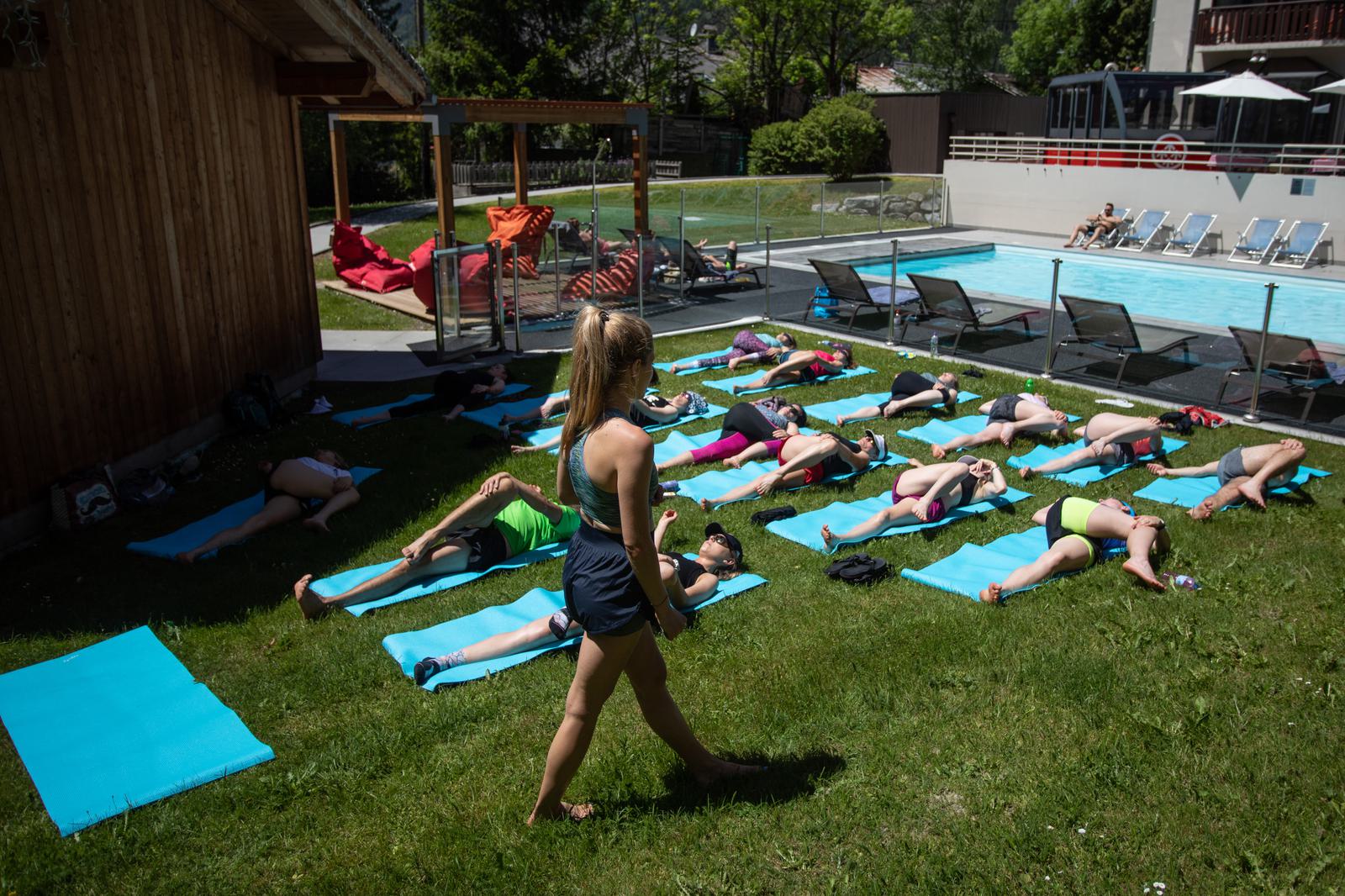 A fitness instructor stands on a grassy area, leading a group of people lying on blue mats performing a stretching exercise outside next to a swimming pool. Other individuals are lounging by the pool and some are seated in a shaded area in the background.