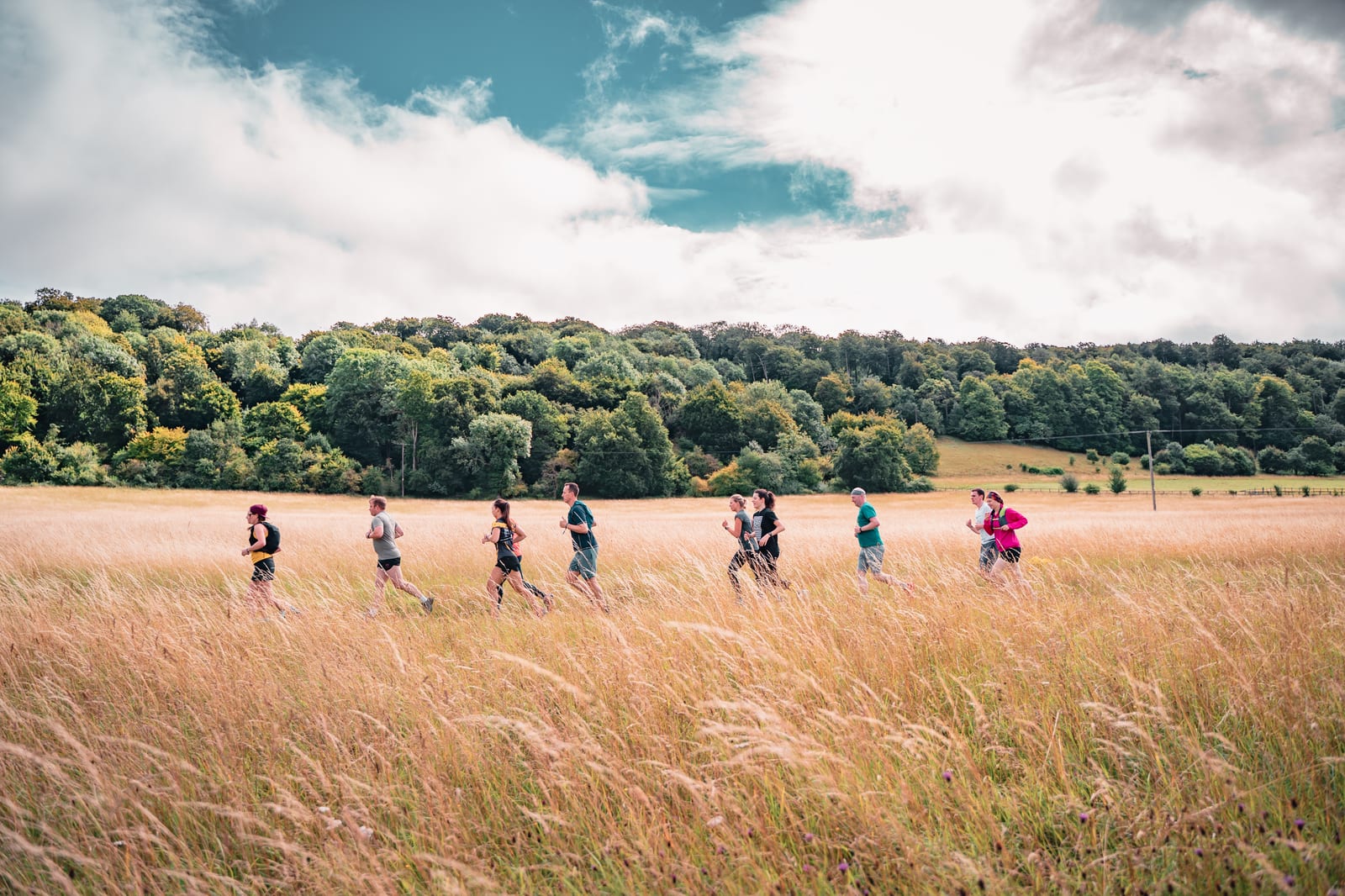 A group of people running through a grassy field with tall, golden grass. The sky is partly cloudy, and there is a dense forest in the background. The runners appear to be enjoying a scenic, outdoor workout.