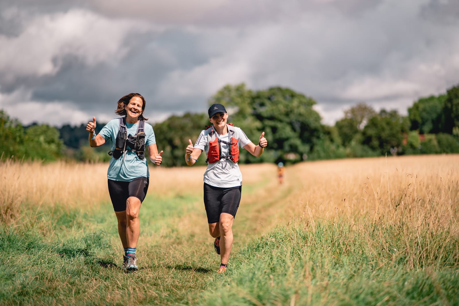 Two women jog on a grassy trail through a wide, open field under a partly cloudy sky. Both are wearing athletic gear and hydration packs. They smile and give thumbs-up signs, enjoying the outdoor activity and serene landscape.