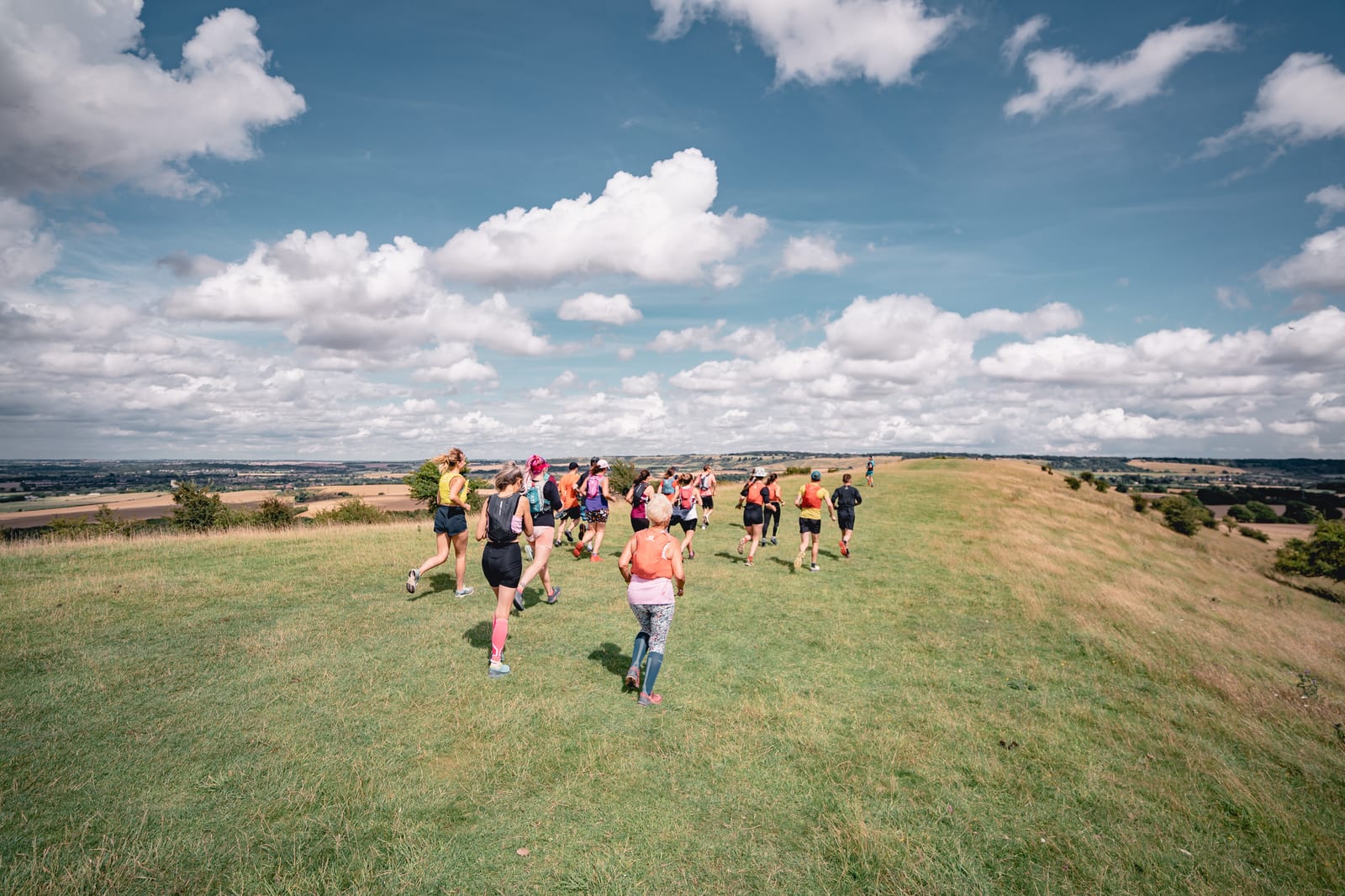 A group of people wearing athletic clothing is running or walking on a grassy hill under a blue sky with scattered clouds. The landscape in the background is expansive, featuring open fields and distant trees. The atmosphere appears lively and energetic.