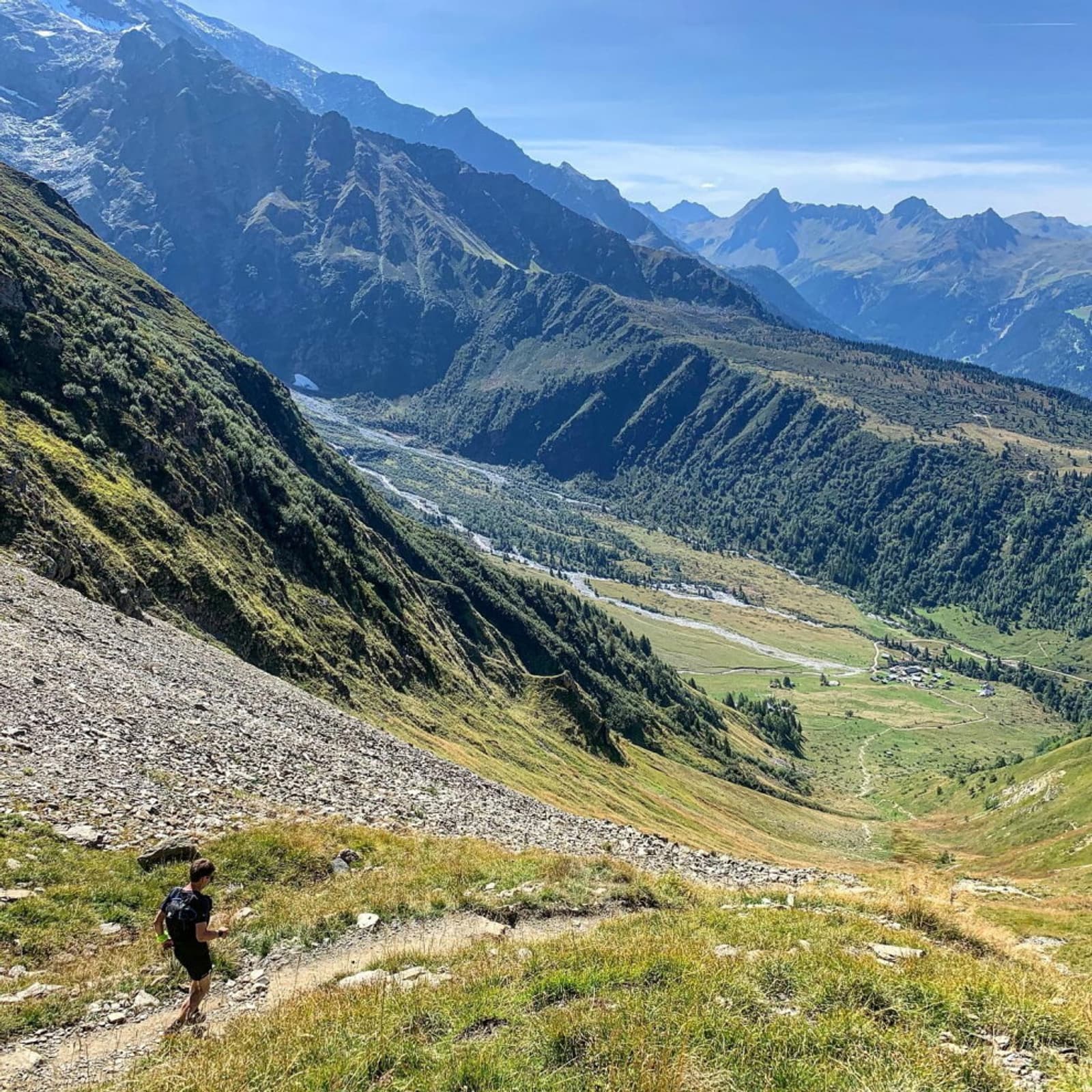A hiker ascends a grassy, rocky mountain path with an expansive view of a valley below, surrounded by lush green slopes and jagged mountain peaks under a clear blue sky.
