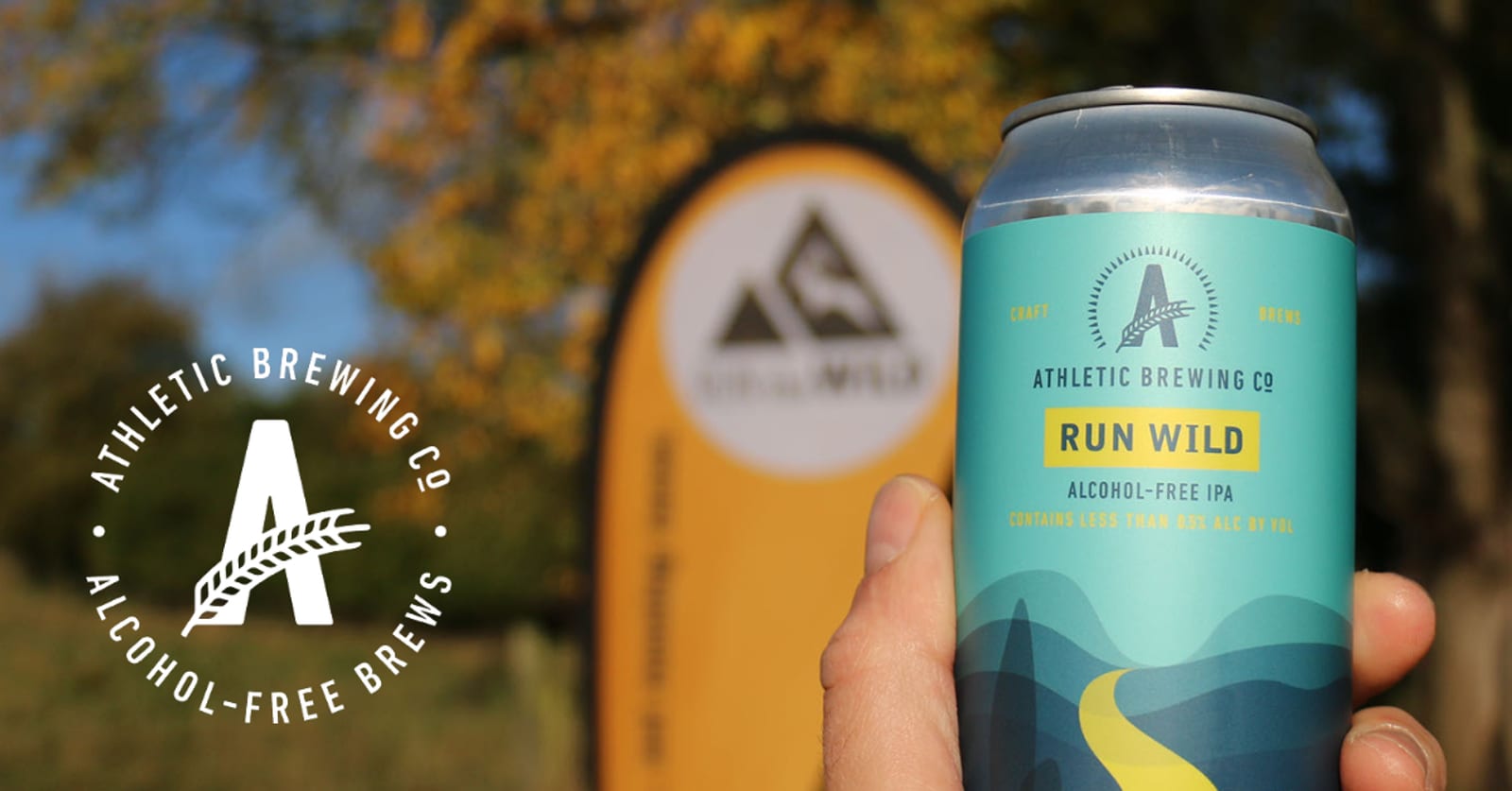 A hand holding a can of Athletic Brewing Co. Run Wild Alcohol-Free IPA. The background shows an outdoor scene with green foliage and a blue sky, along with a large Athletic Brewing Co. sign. The logo and slogan are also visible on the left side of the image.
