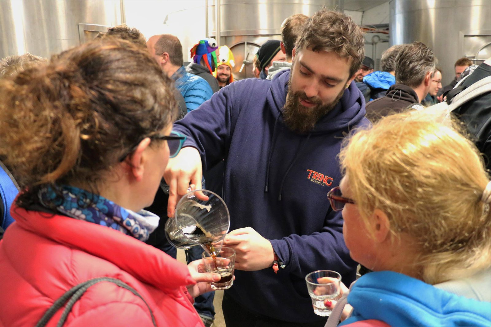 A bearded man in a navy blue hoodie serves beverages to two women in colorful jackets at a crowded event. The venue appears to be a brewery or similar setting, with large metal tanks in the background. Several other people are also gathered around.