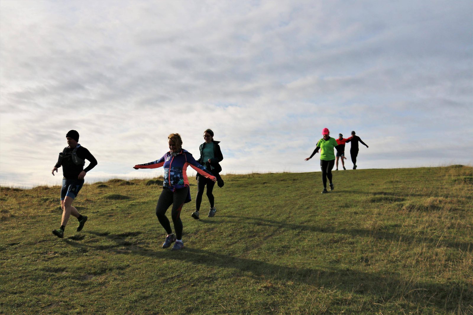 A group of people are running downhill on a grassy hill under a cloudy sky. They are dressed in athletic gear, with some wearing hats and gloves. The mood appears joyful and energetic as they descend the slope.