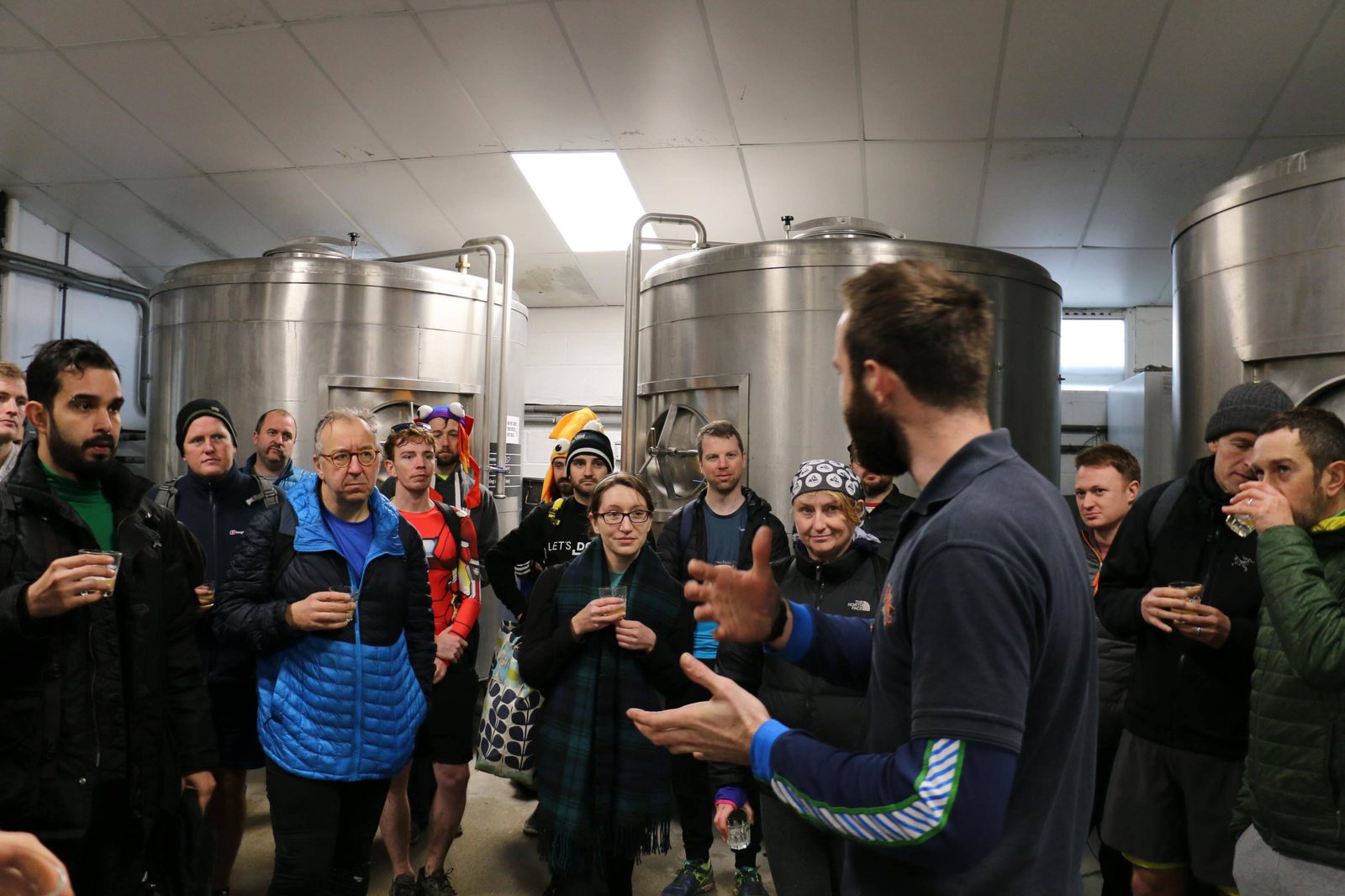 A group of people attentively listen to a speaker inside a brewery, surrounded by large stainless steel tanks. Many hold small cups and wear casual outdoor clothing. The atmosphere suggests an informative tour or presentation.