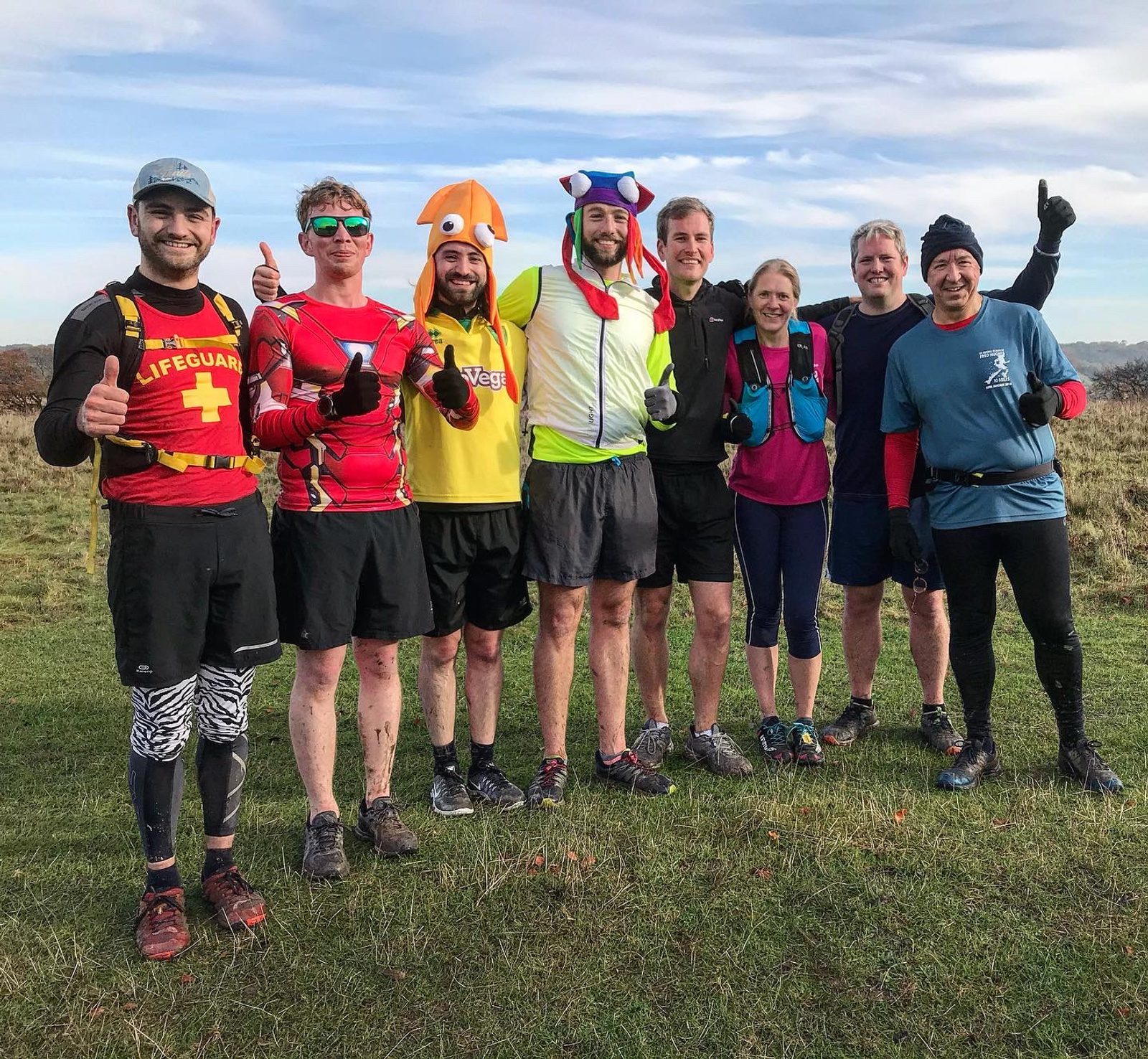 A group of eight people standing on a grassy field, dressed in various running outfits. Some wear costumes, including a lifeguard outfit and a colorful hat. They are smiling and giving thumbs up, with a clear sky and trees in the background.