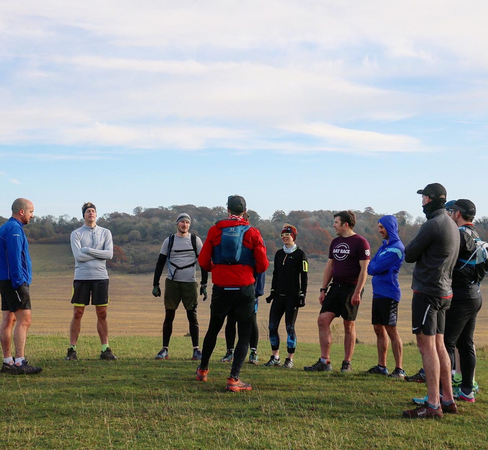A group of people dressed in athletic clothing stand in a circle on a grassy field, with some wearing hats and backpacks. The sky is partly cloudy, and trees are visible in the background. The group appears to be preparing for a run or outdoor activity.