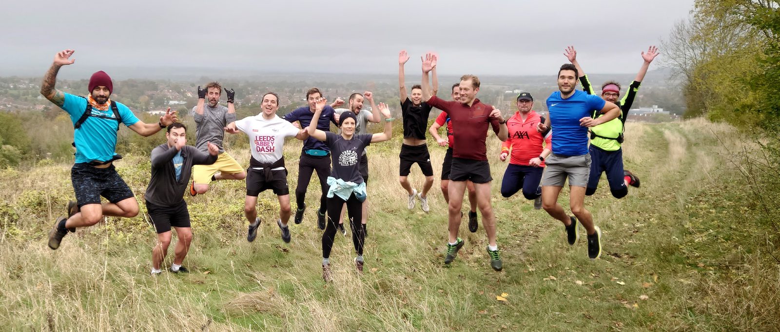 A group of twelve people are outdoors on a grassy trail, jumping in the air with their arms raised. They are dressed in athletic gear with a mix of shorts, leggings, and jackets. The background shows a partially cloudy sky and a distant view of trees and buildings.