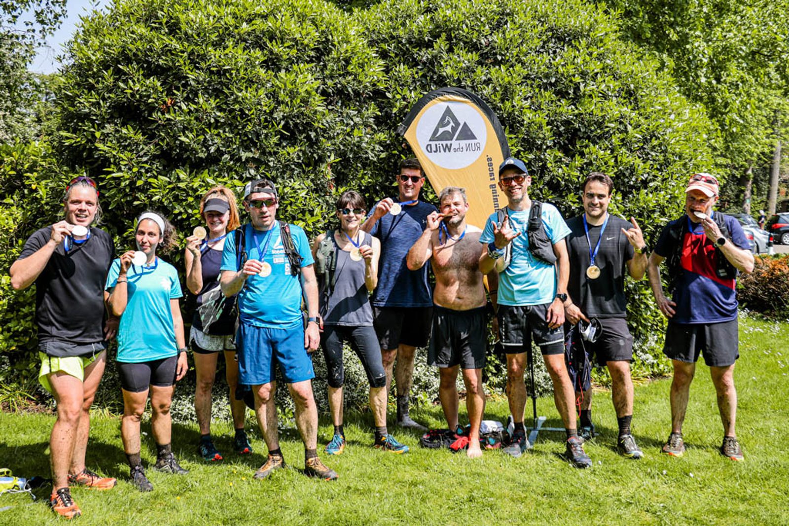 A group of eleven runners, wearing athletic gear and medals, pose together for a photo in front of a green hedge. They appear sweaty and tired but happy, holding up their medals with proud expressions. A race event sign is visible in the background.