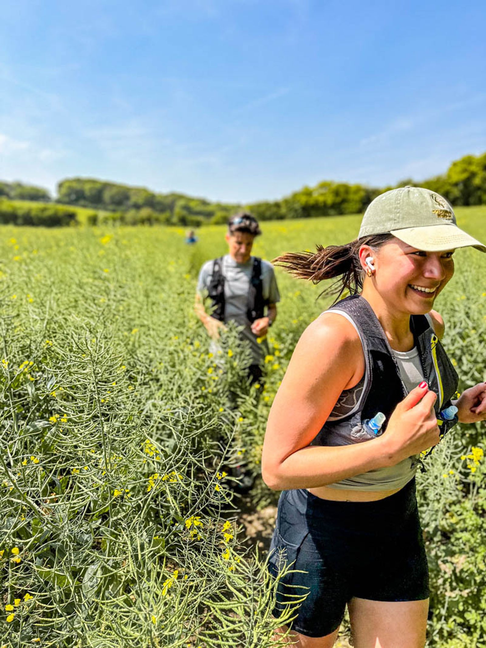 Two people are running through a field of yellow flowering plants on a sunny day. The person in the foreground, wearing a gray cap, black sports bra, and black shorts, is smiling. The second person, slightly behind and blurred, is also running.