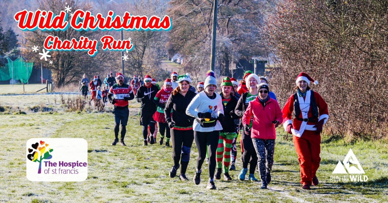 A group of people in festive costumes, including Santa outfits and elf hats, run outdoors on a frosty path during the “Wild Christmas Charity Run” for The Hospice of St Francis. Bright sunlight and winter trees are in the background.