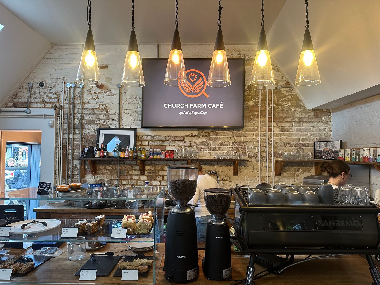 A cozy café interior with exposed brick walls, hanging lights, a counter displaying various pastries, coffee grinders, and an espresso machine. Two staff members work behind the counter beneath a sign reading "Church Farm Café.