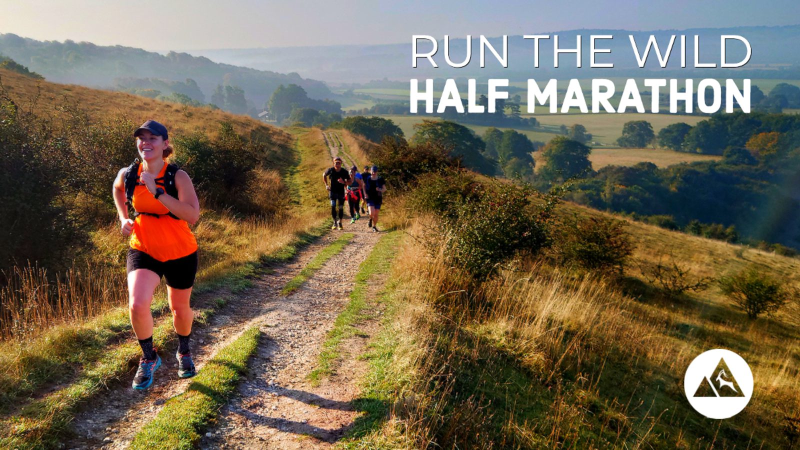 A group of runners participate in the "Run the Wild Half Marathon" on a scenic trail surrounded by rolling hills and trees. The lead runner, wearing an orange top and cap, smiles while running under a clear sky. The event logo is visible in the bottom right corner.