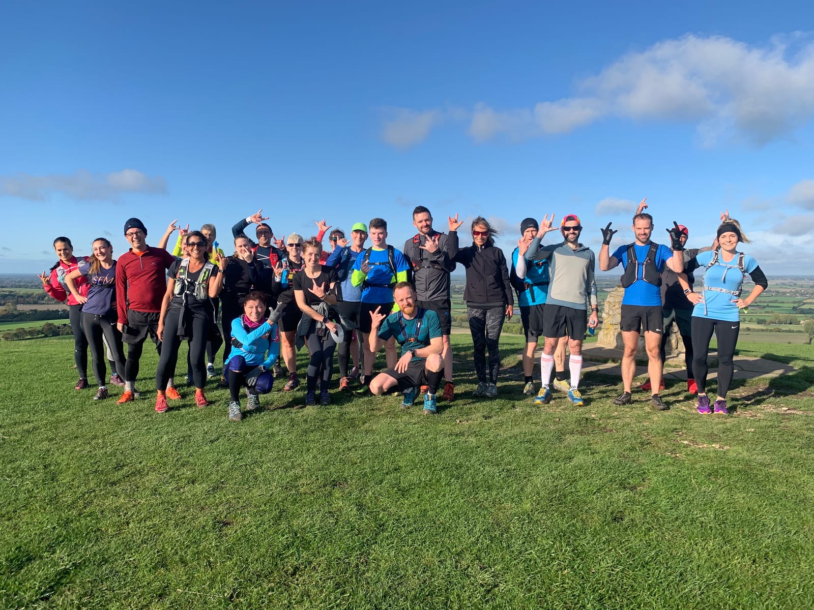 A group of people standing together outside on a grassy hilltop under a clear blue sky. They are wearing athletic clothing and appear to be happy and energetic. Many are posing with playful gestures, and there is a scenic landscape in the background.