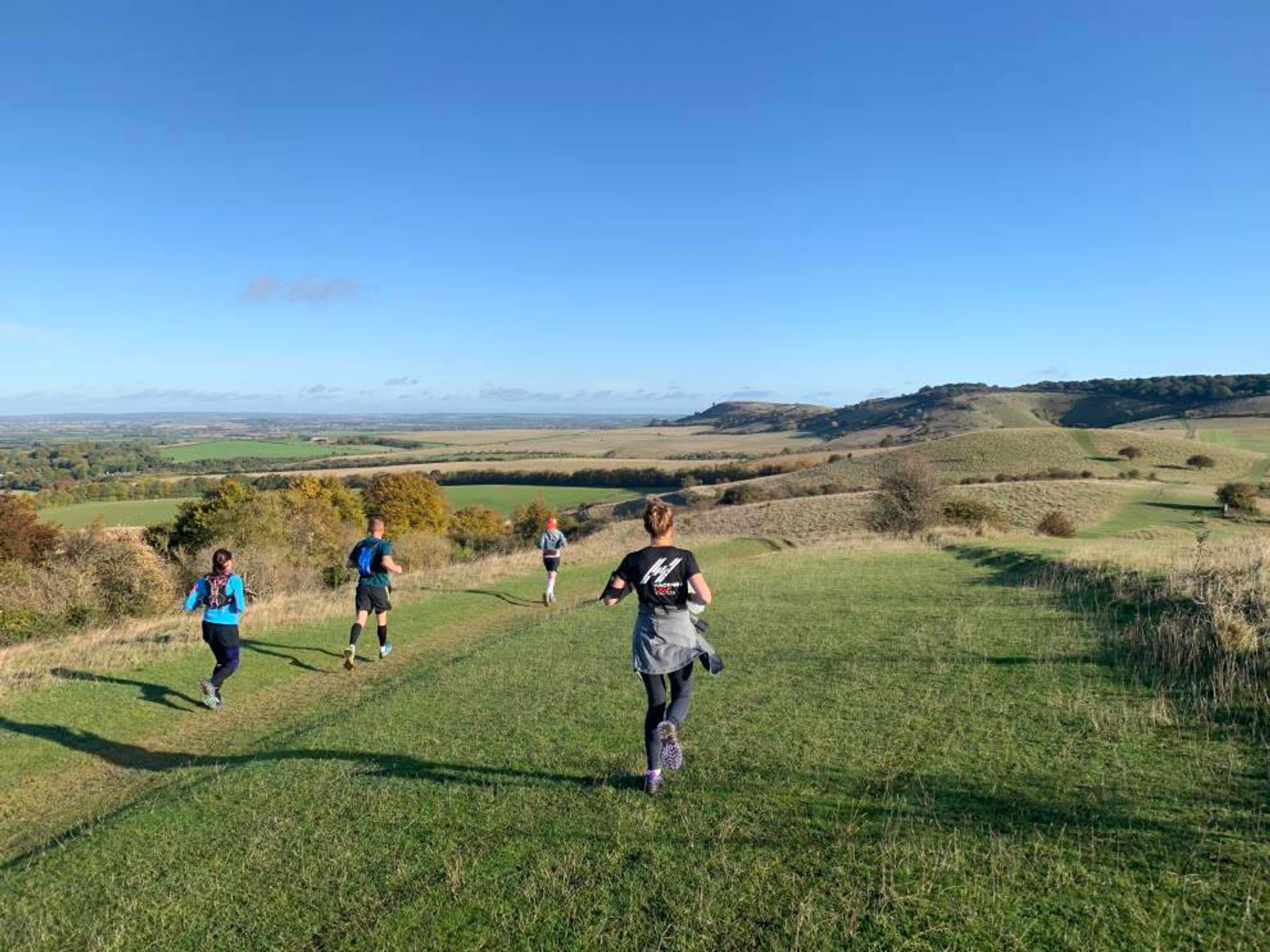 A group of people are jogging along a grassy path on a hill under a clear blue sky. The landscape features rolling hills, scattered trees, and expansive fields. One jogger is in the foreground wearing a gray top and purple leggings, with others further along the path.