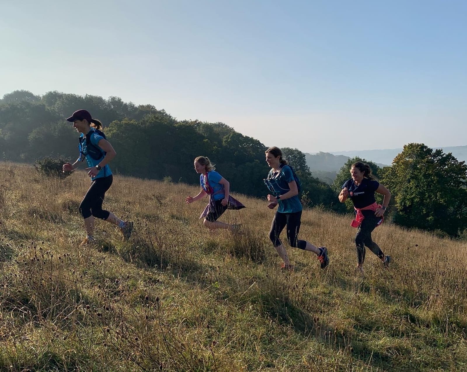Four people are running uphill on a grassy field in the early morning. They are dressed in athletic wear and seem focused. The background features a tree line under a clear sky. The light suggests it is shortly after sunrise, casting a soft glow on the scene.