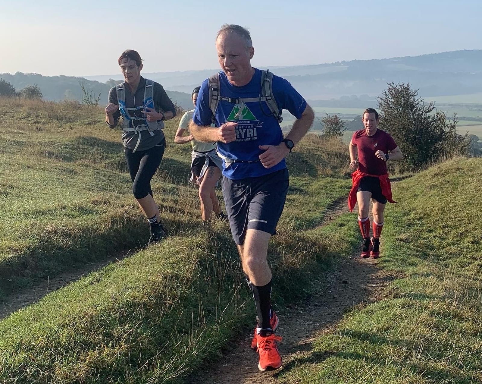 Four people are running up a grassy hill, with a scenic landscape and hazy mountains in the background. The lead runner, wearing a blue shirt and orange shoes, looks focused. The other runners follow closely, each at their own pace. The weather appears clear and sunny.