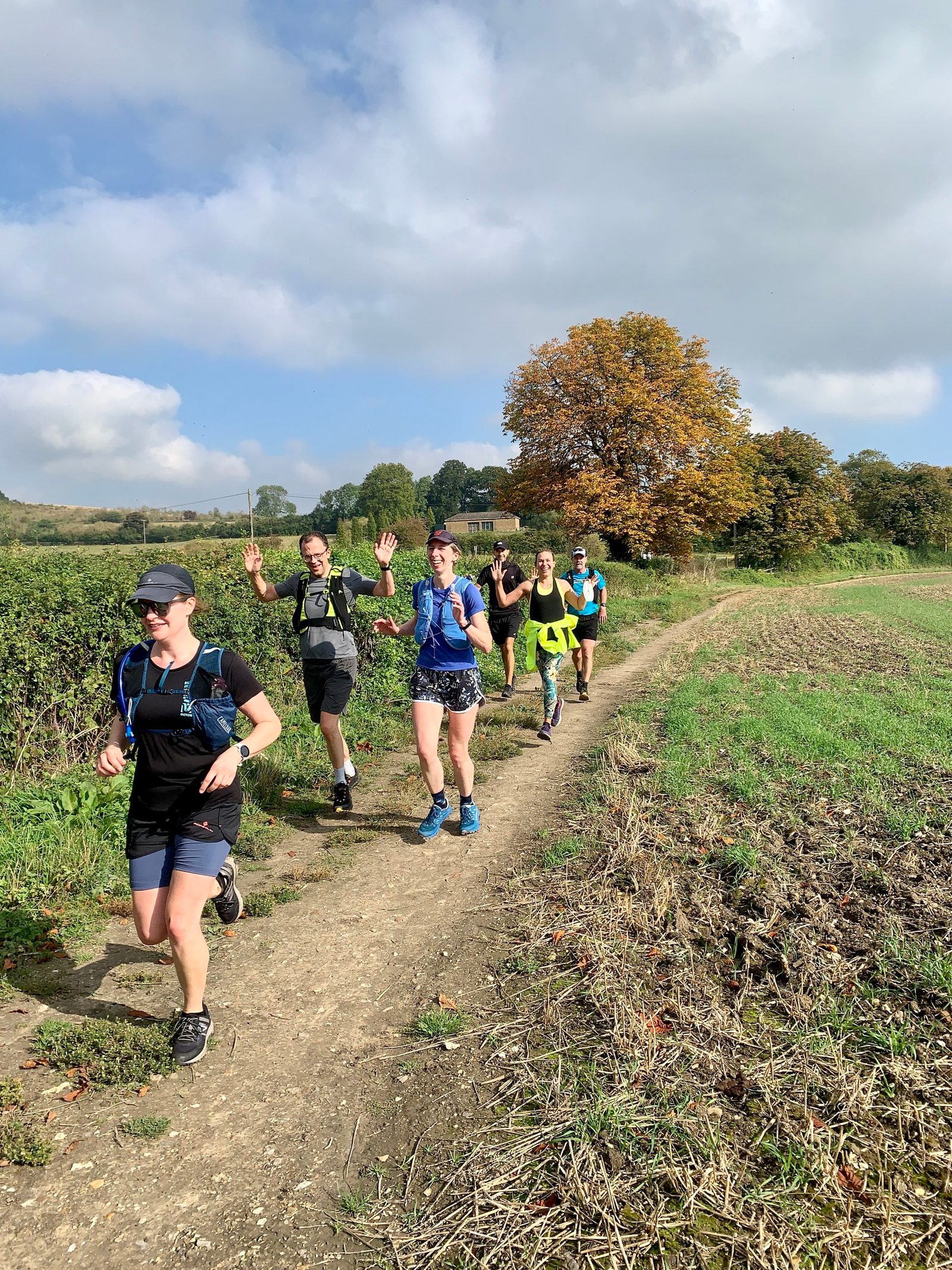 A group of six people in athletic wear are running on a dirt trail through a rural landscape. They appear happy and some are smiling, with one person waving at the camera. The background features green fields and autumn trees under a partly cloudy sky.