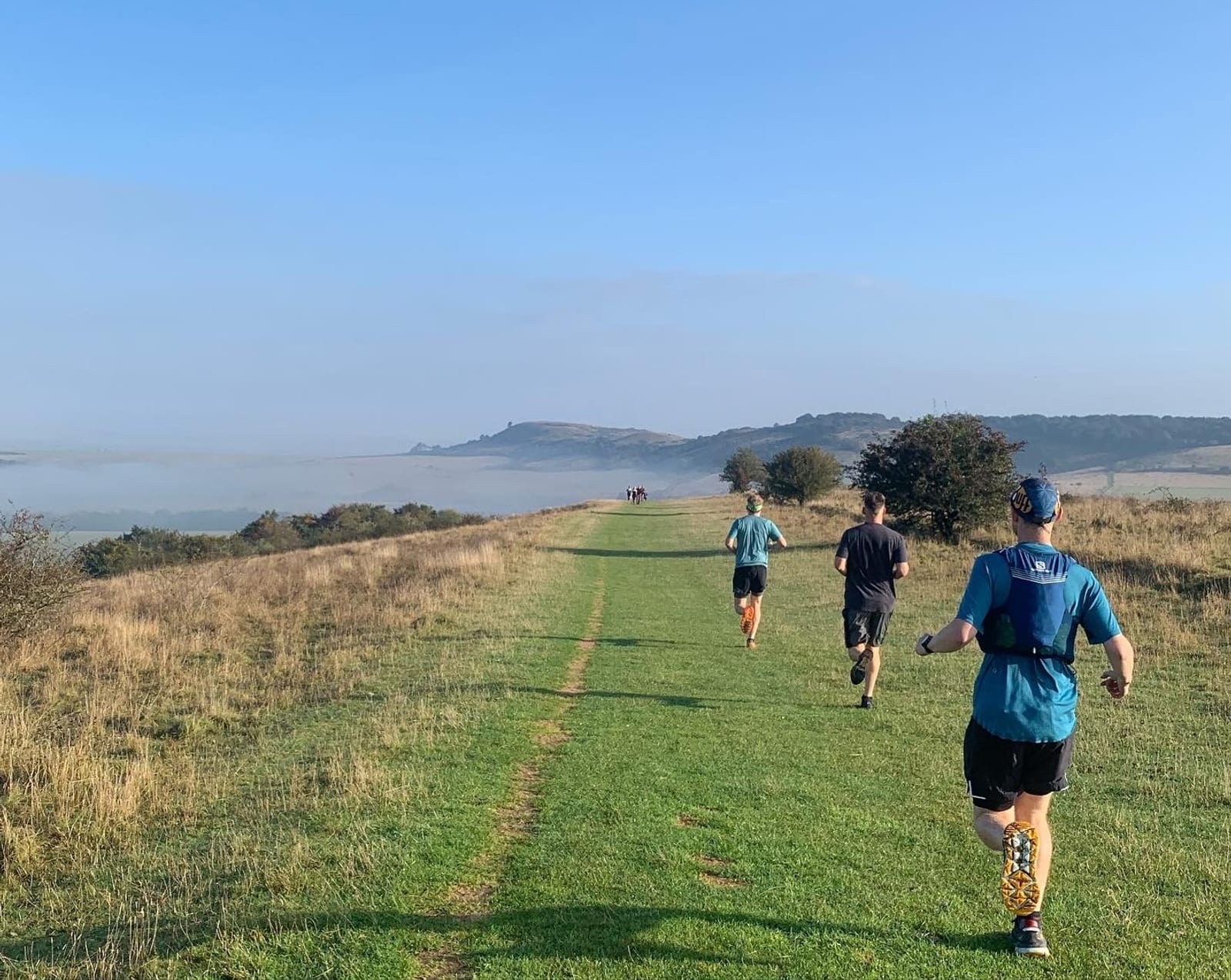 Three people are running on a grassy trail atop a hilly landscape under a clear blue sky. The path is flanked by patches of dry grass and scattered bushes. In the background, there are distant hills and other runners further along the trail.